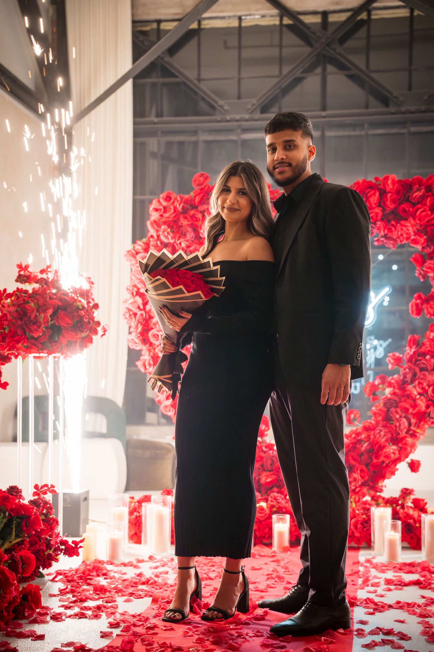 Romantic scene with sparklers, red rose wall, and couple in formal attire.