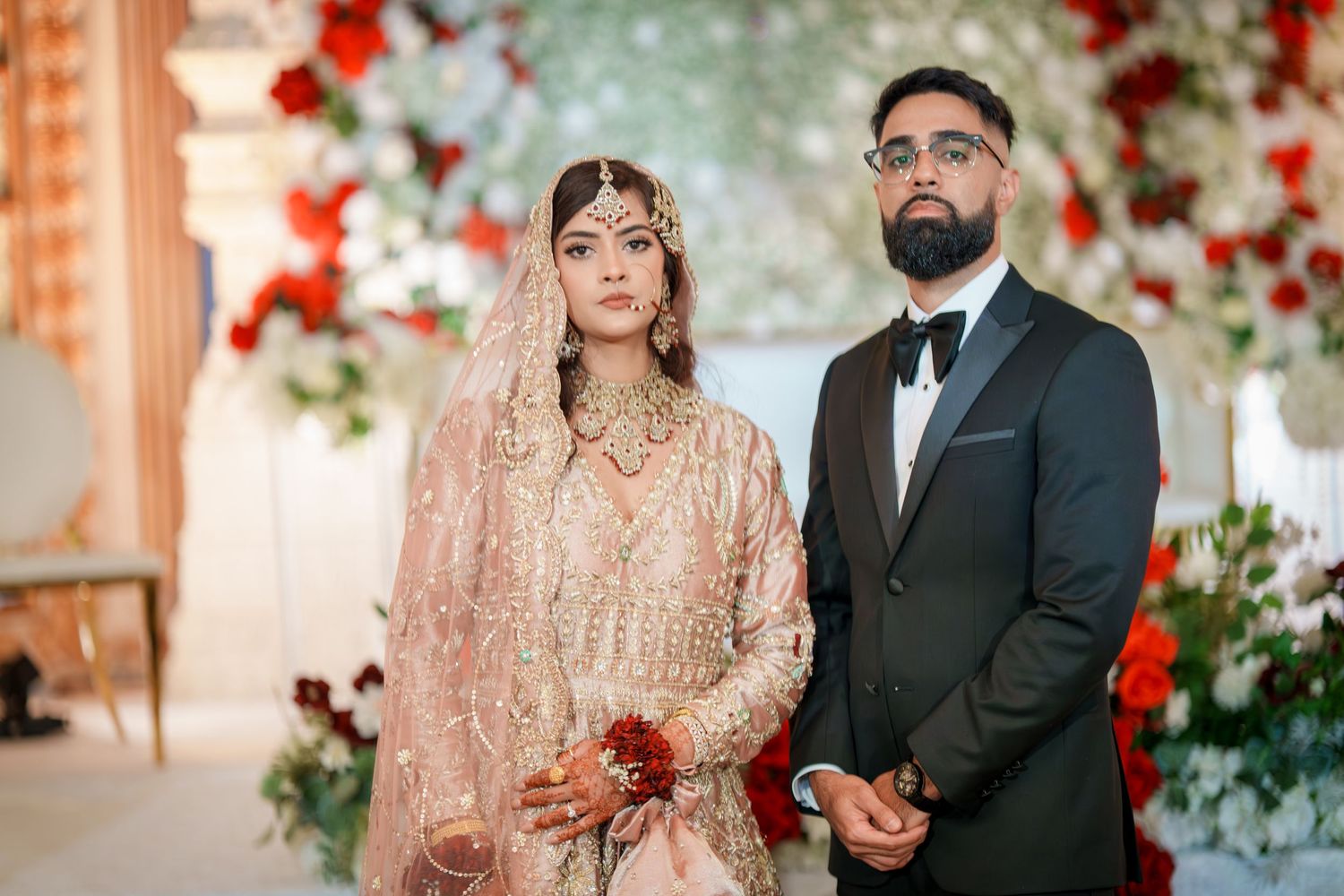 Wedding couple in traditional South Asian attire posing against a backdrop with red floral decorations.