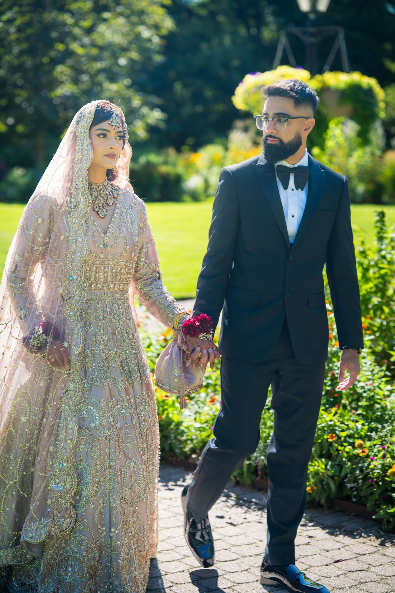 A couple in traditional South Asian wedding attire walking outdoors, with the bride in a detailed peach lehenga.
