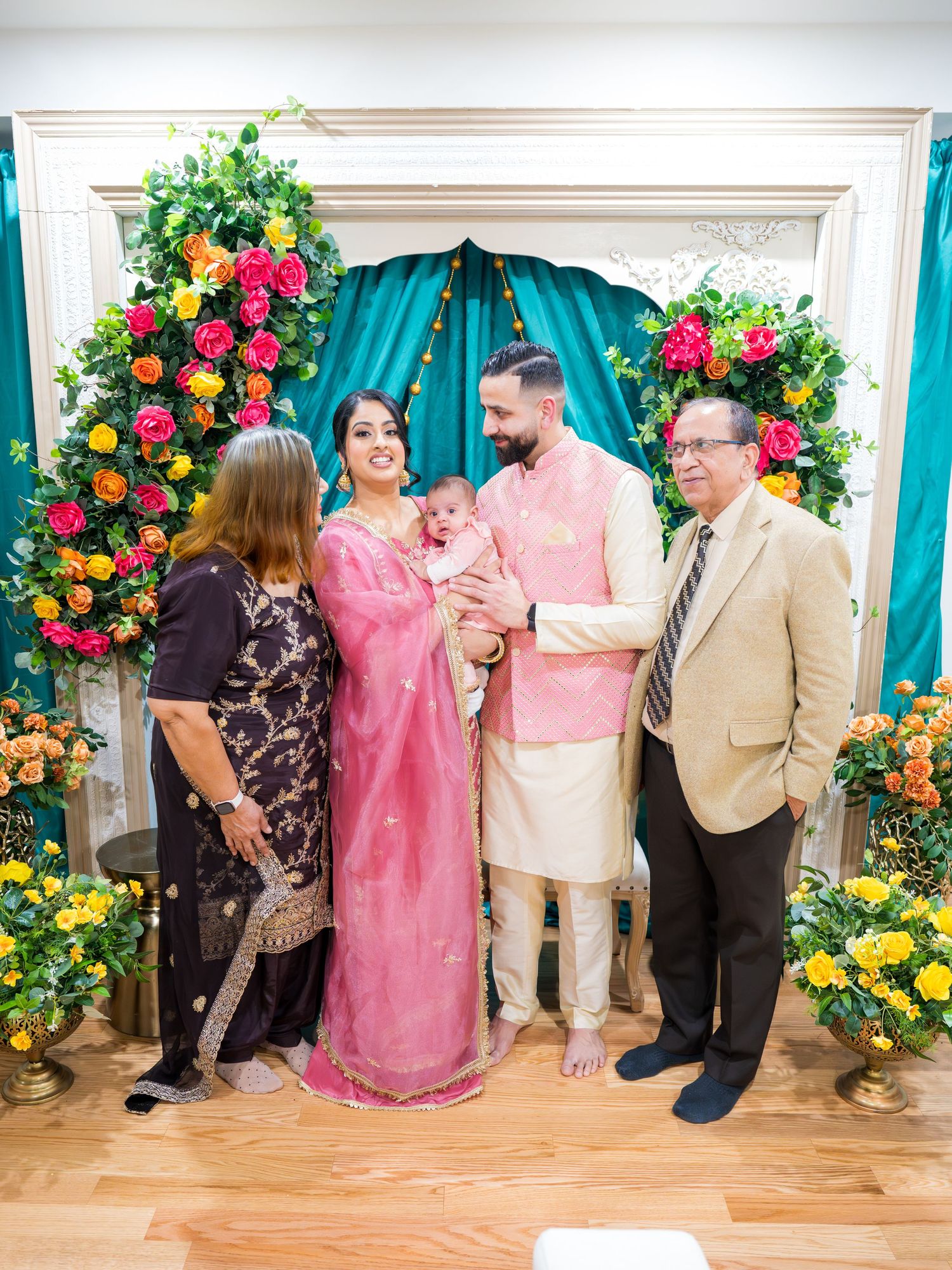 Family of five posing with newborn at colorful floral backdrop during Indian naming ceremony.