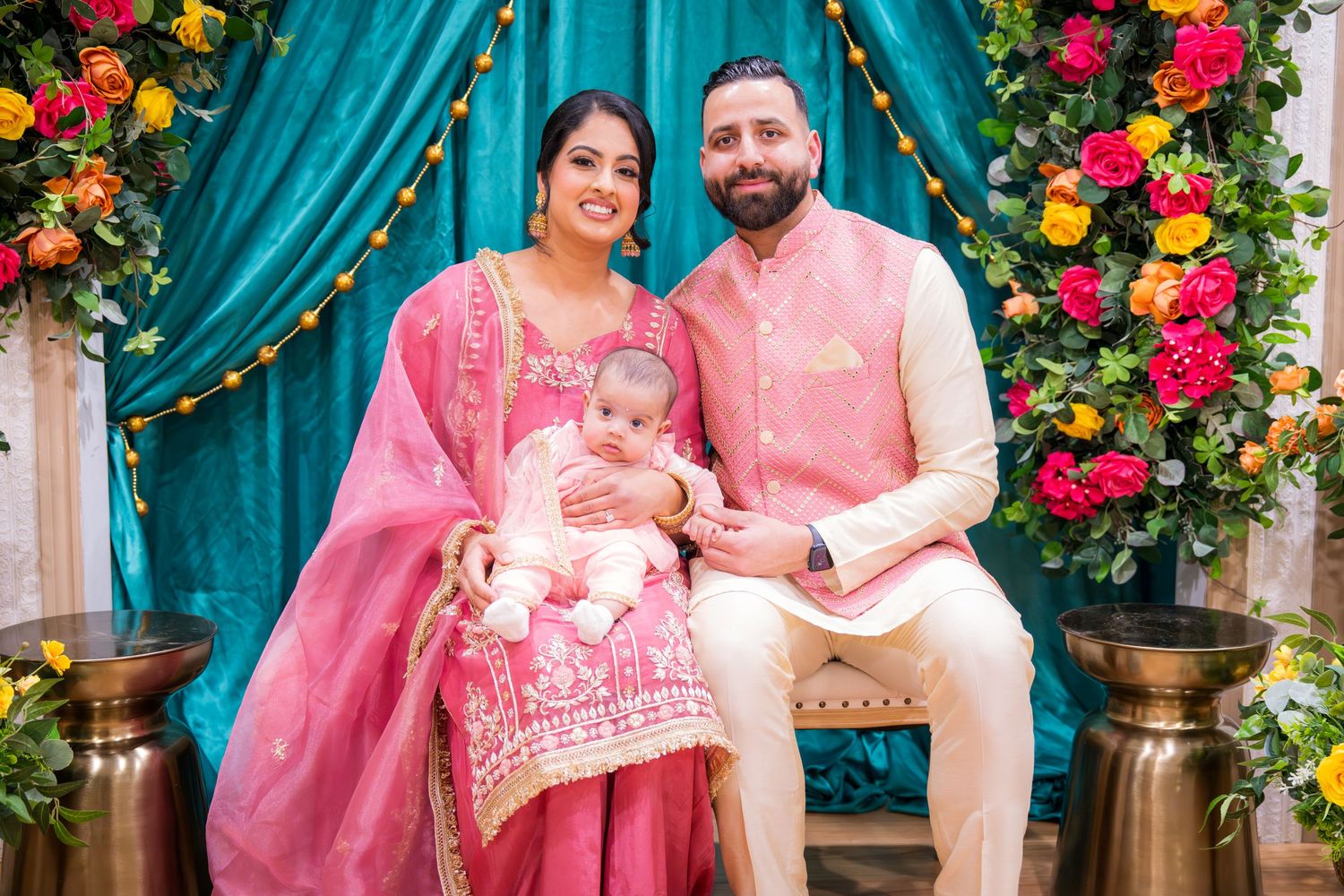 South Asian family in pink traditional Indian attire posing with baby at decorated ceremonial event.