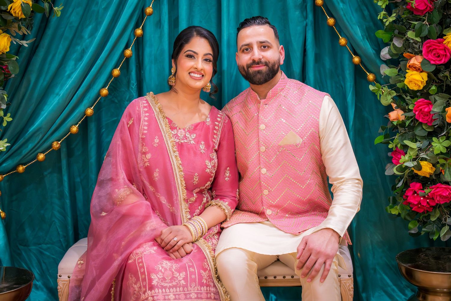 South Asian couple in matching pink traditional Indian outfits seated at floral ceremony backdrop.