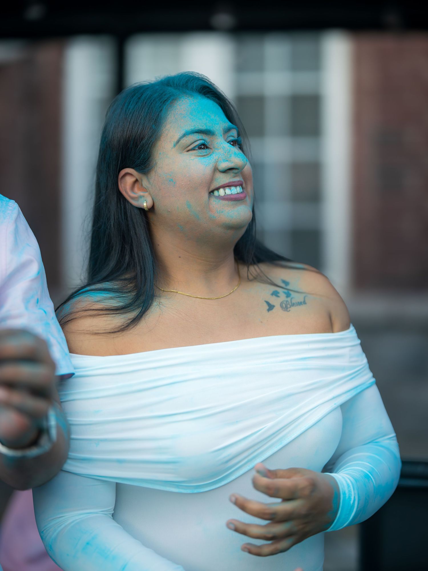 A smiling woman in a white off-shoulder dress with blue powder paint on her face at a Holi celebration.