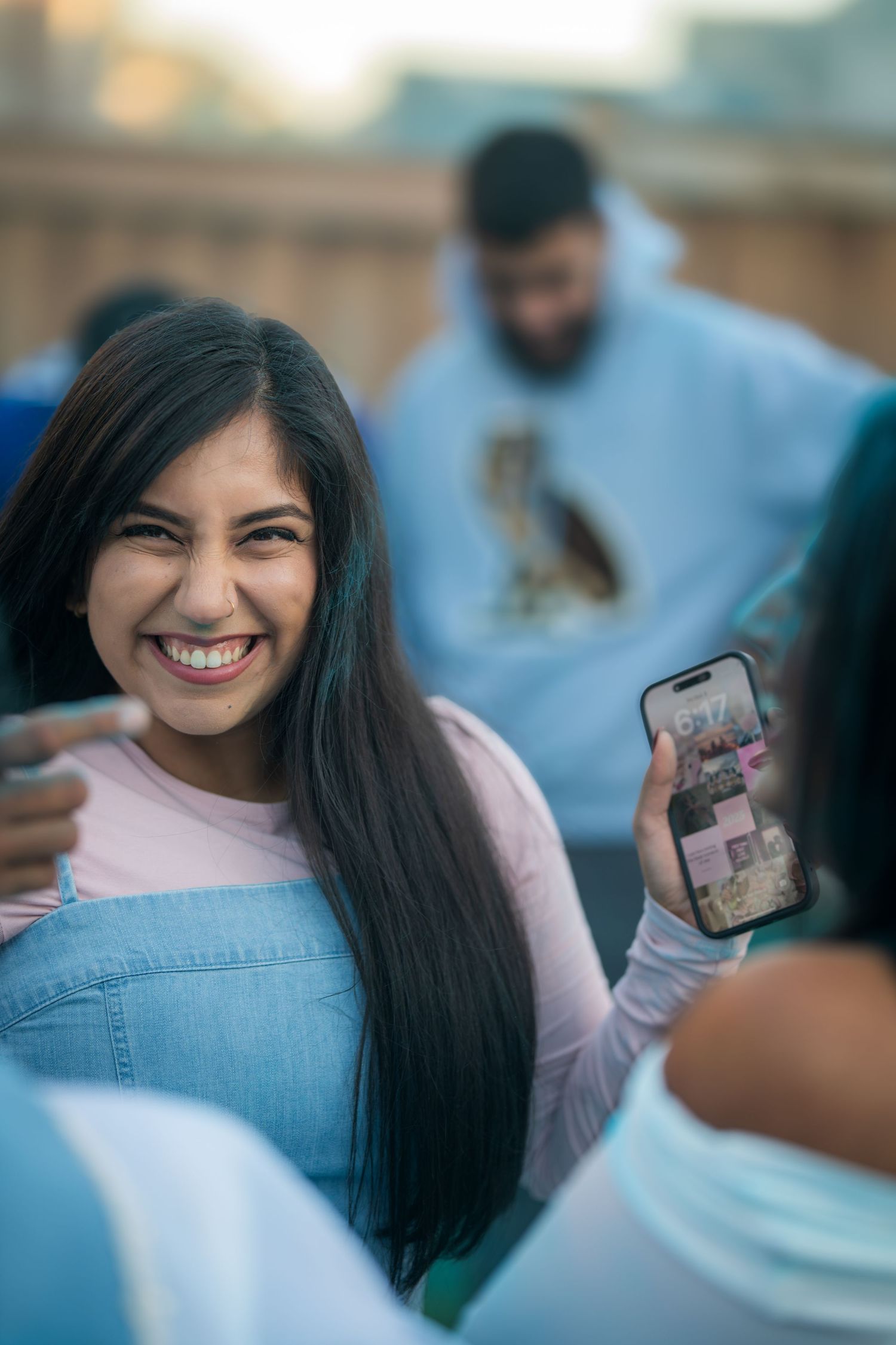 Laughing young woman in denim overalls holds phone at an outdoor gender reveal party.