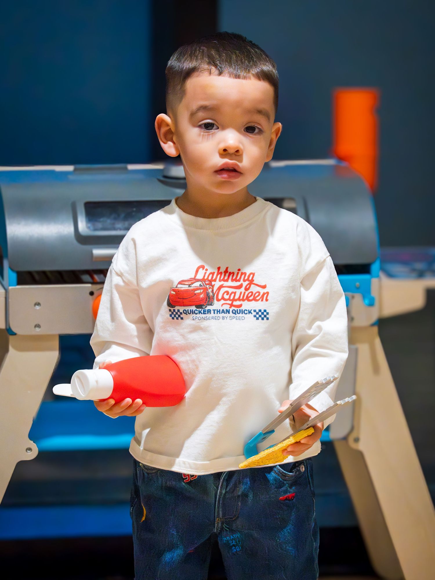 Young toddler boy in Lightning McQueen shirt holds a red bottle while playing at an indoor activity center.