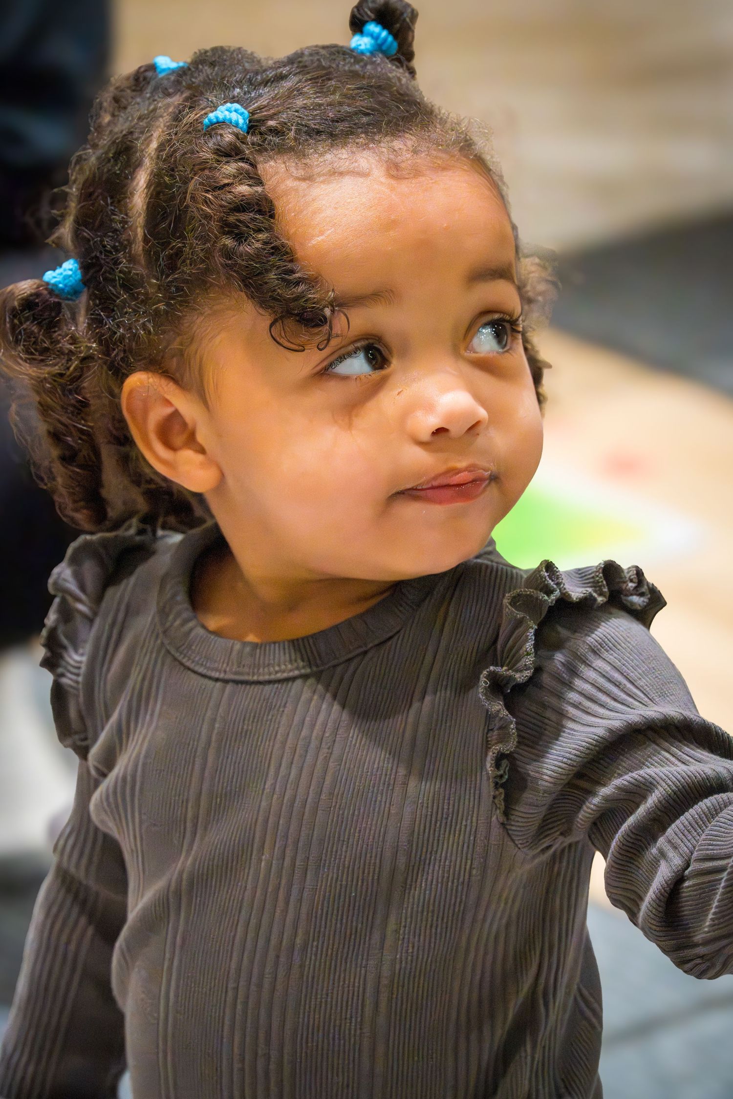 Adorable young girl with blue hair ties wearing a dark ribbed long-sleeve top, looking up curiously.