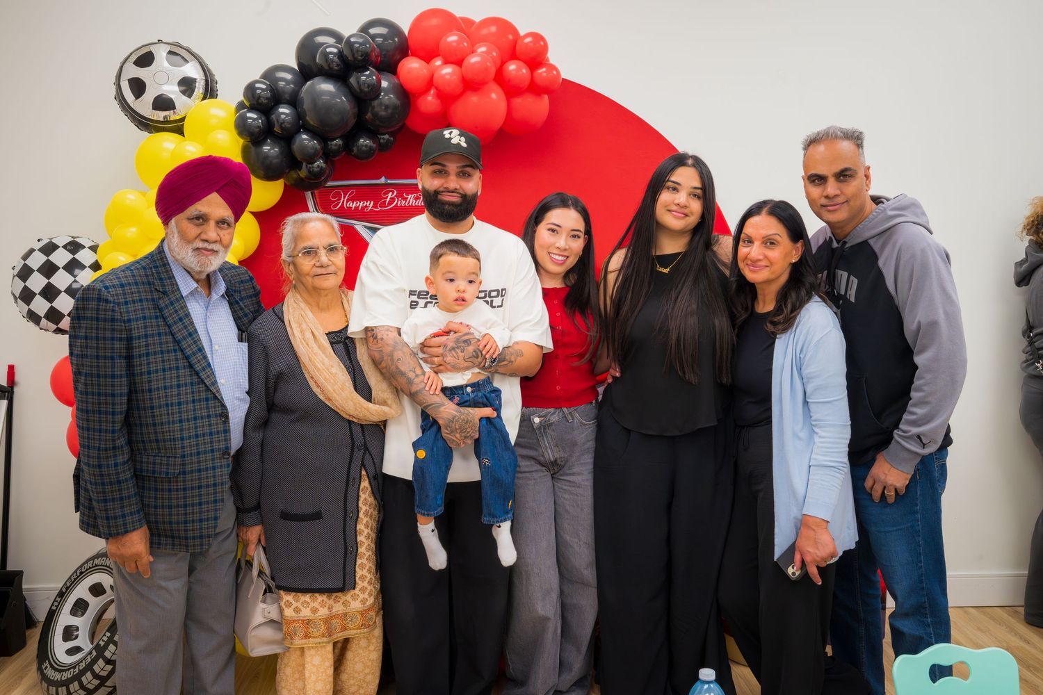 A multigenerational family group of seven posing together at a colorful birthday party with red, yellow, and black balloons.