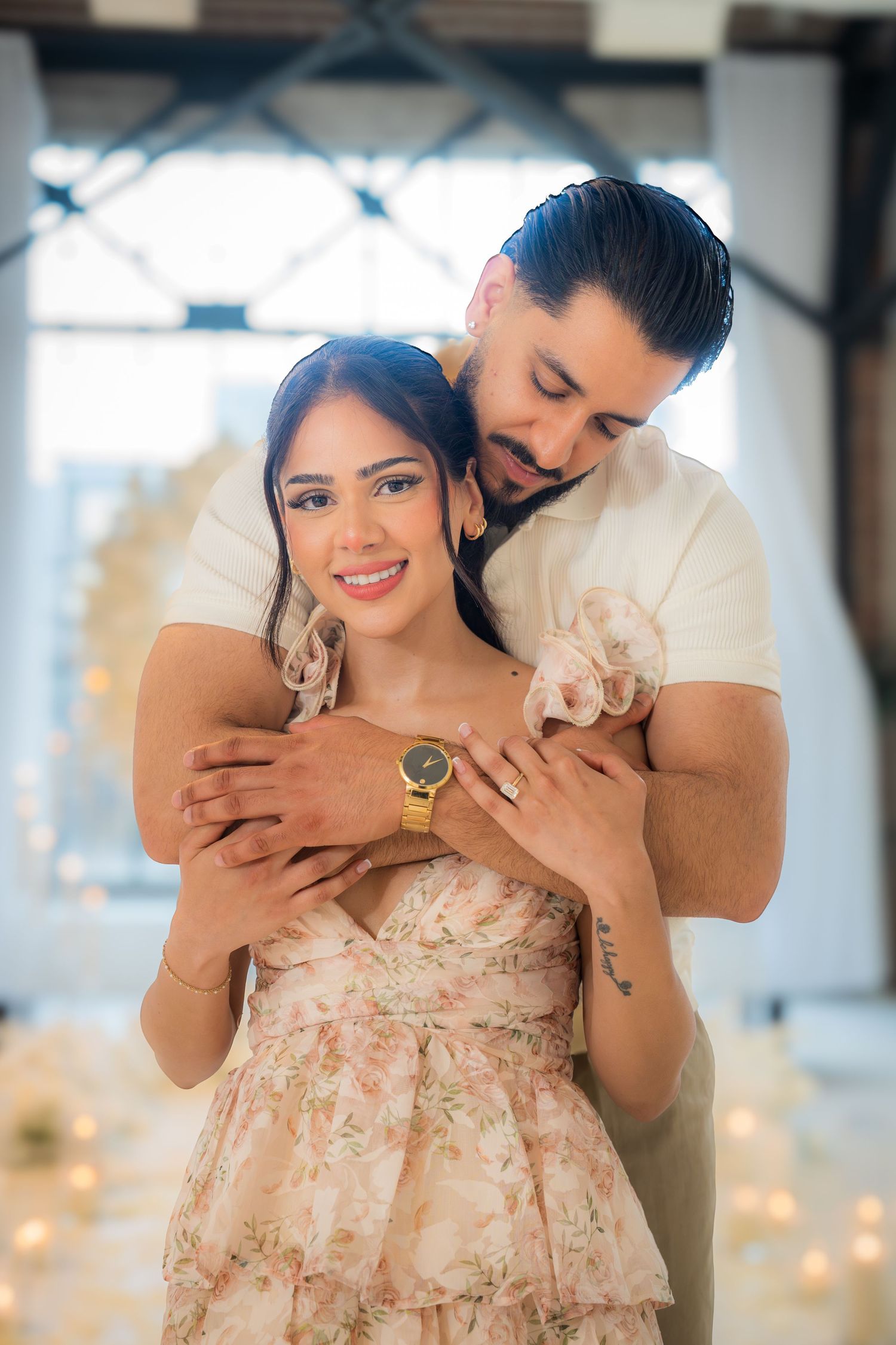 Man embracing woman from behind as she smiles, both wearing matching gold bracelets in a romantic setting.