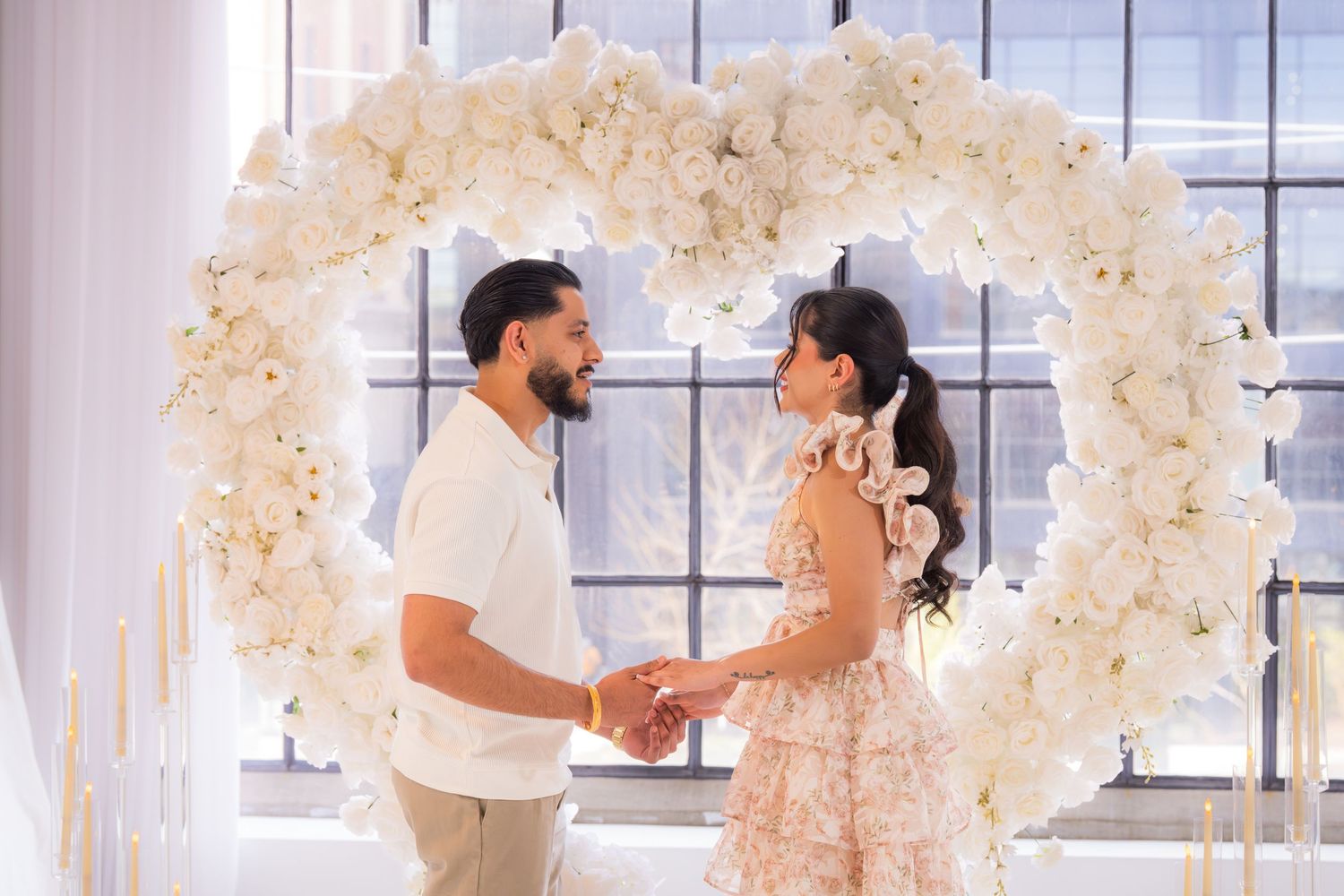 Happy couple holding hands and gazing at each other under a stunning white floral heart-shaped arch with soft lighting.