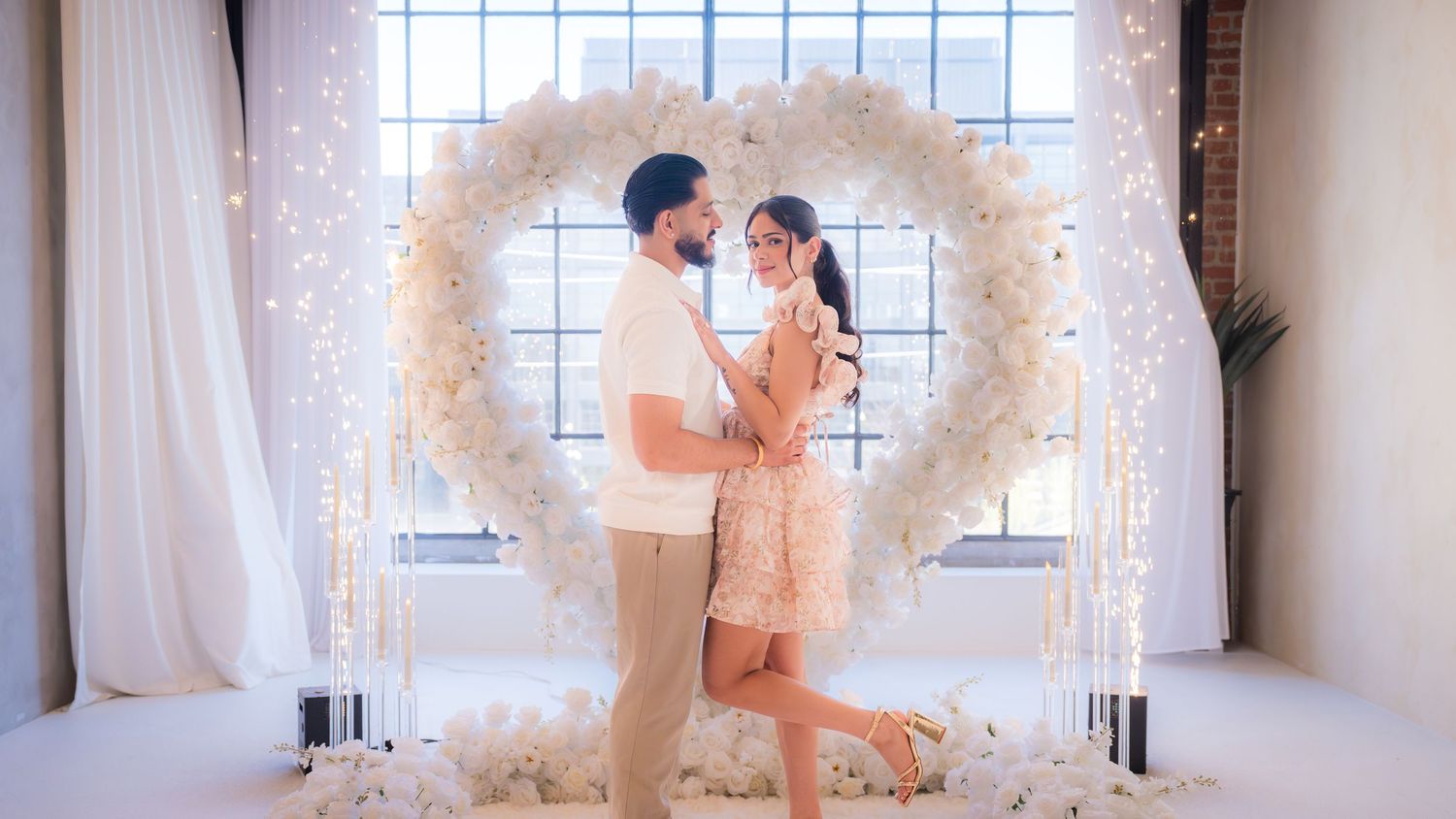 Couple sharing a tender moment in front of a white floral heart arch with fairy lights.