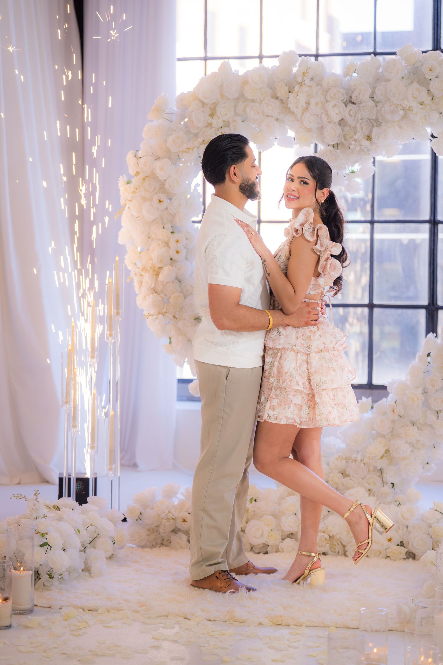 Couple standing under white floral heart arch with sparkling fireworks display.