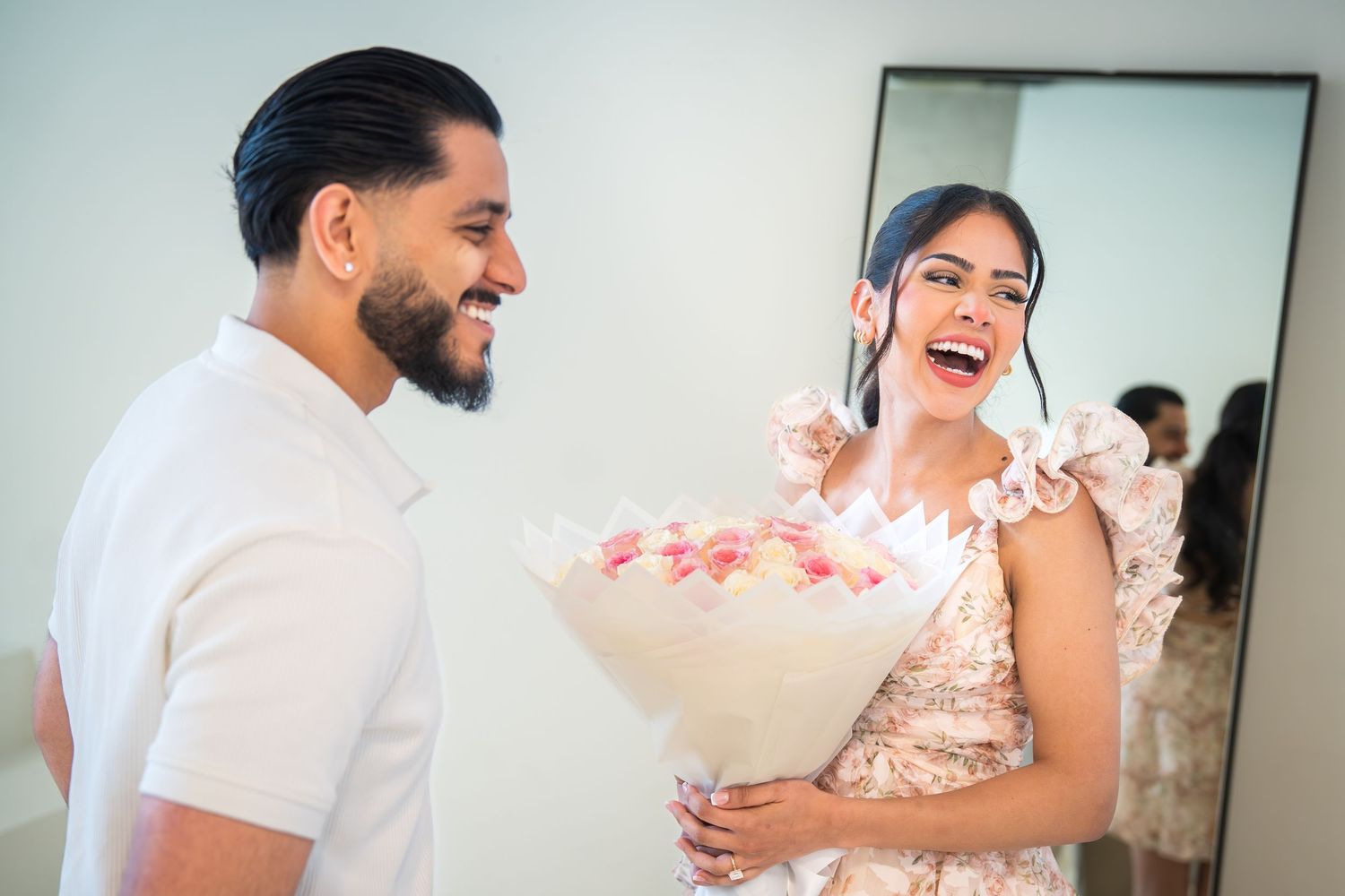 A man presents a pink flower bouquet to a laughing woman in a floral dress near a mirror.