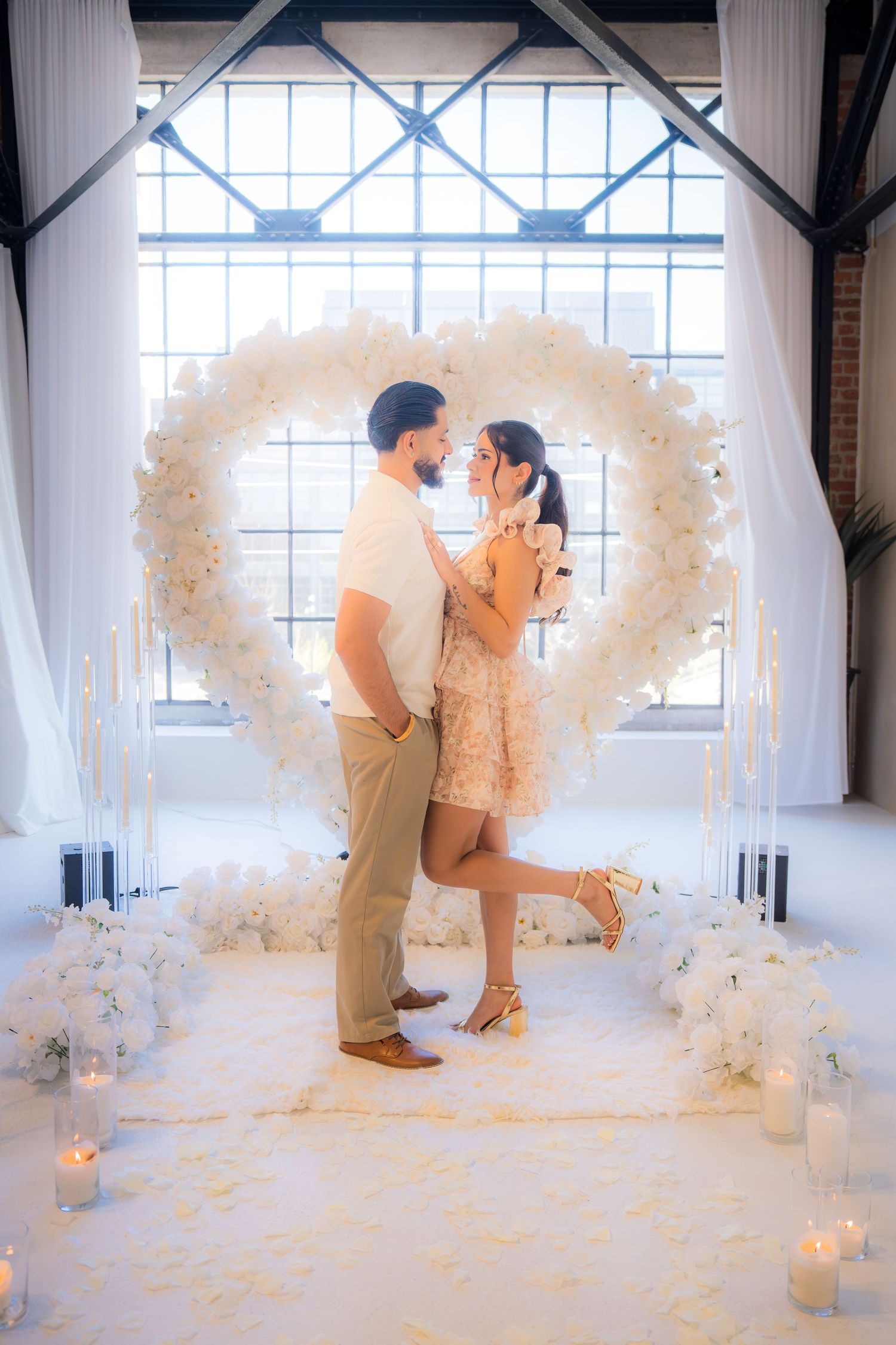 An engaged couple embraces in front of a large white floral heart arch with candles and flowers.