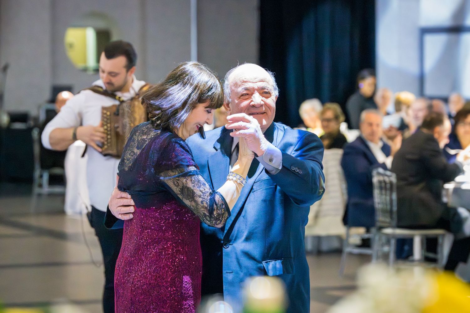Elderly man in blue suit dancing with woman in purple dress at wedding reception celebration.