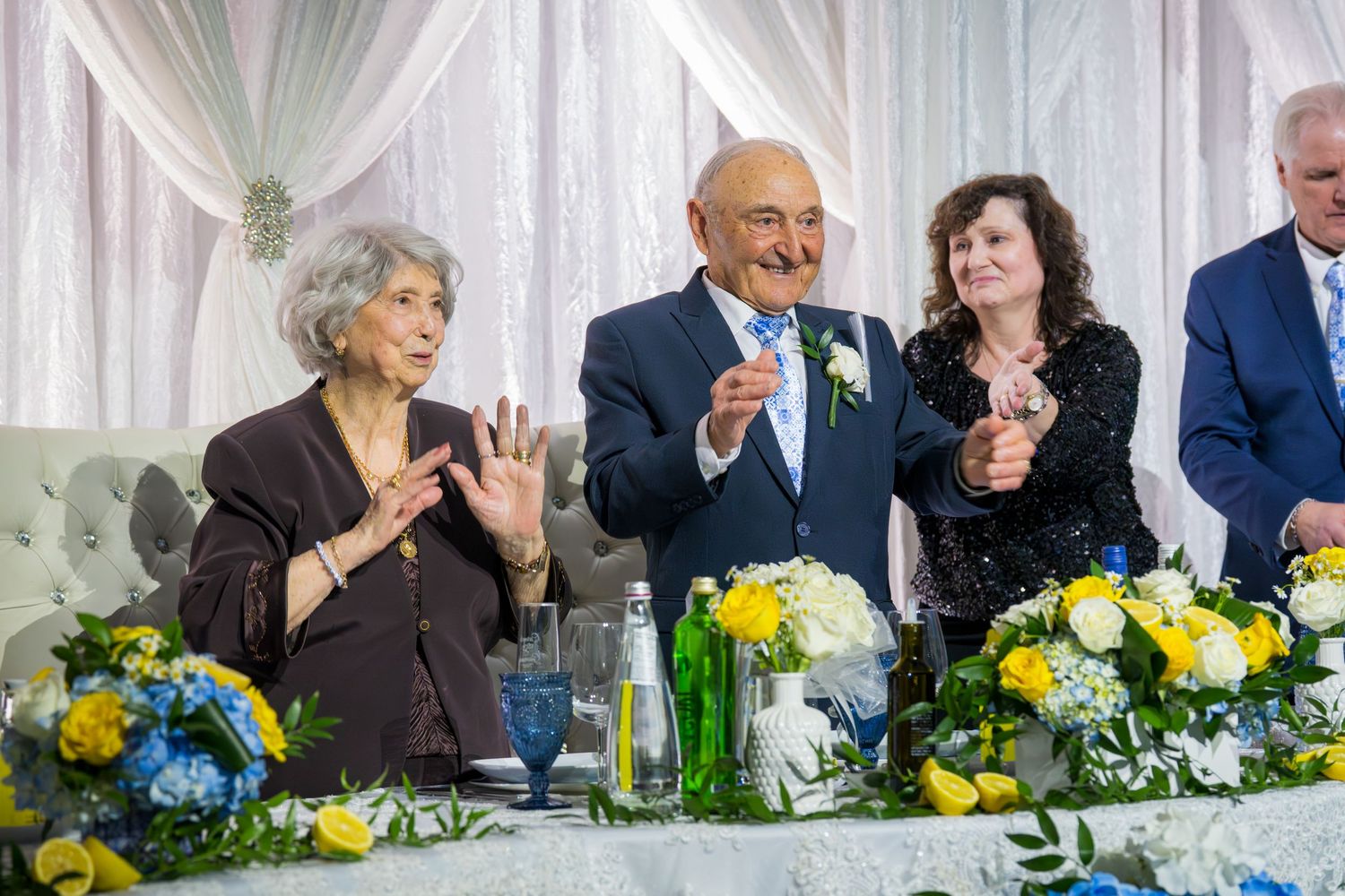 Elderly couple and woman celebrate at a wedding reception with yellow and blue floral arrangements on the table.