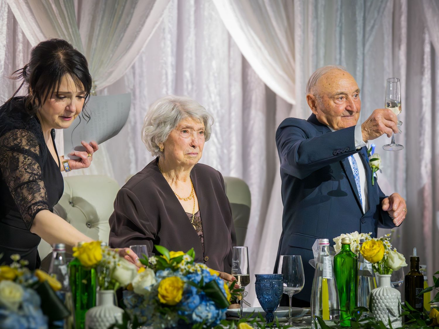Elderly man raising champagne glass for toast at anniversary celebration dinner with family seated at decorated table.