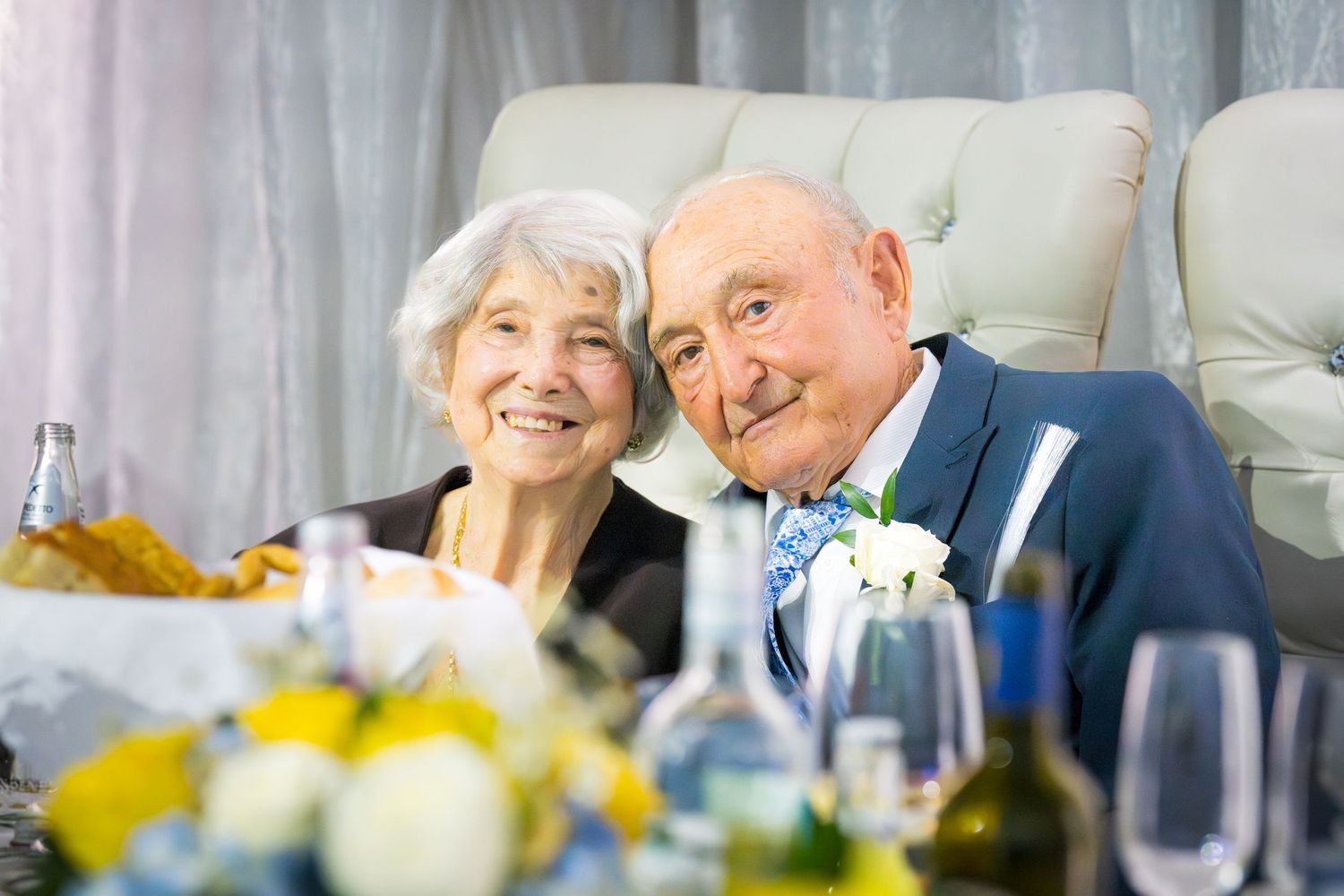 Happy elderly couple smiling together at a celebratory dinner table with flowers and glasses in the foreground.