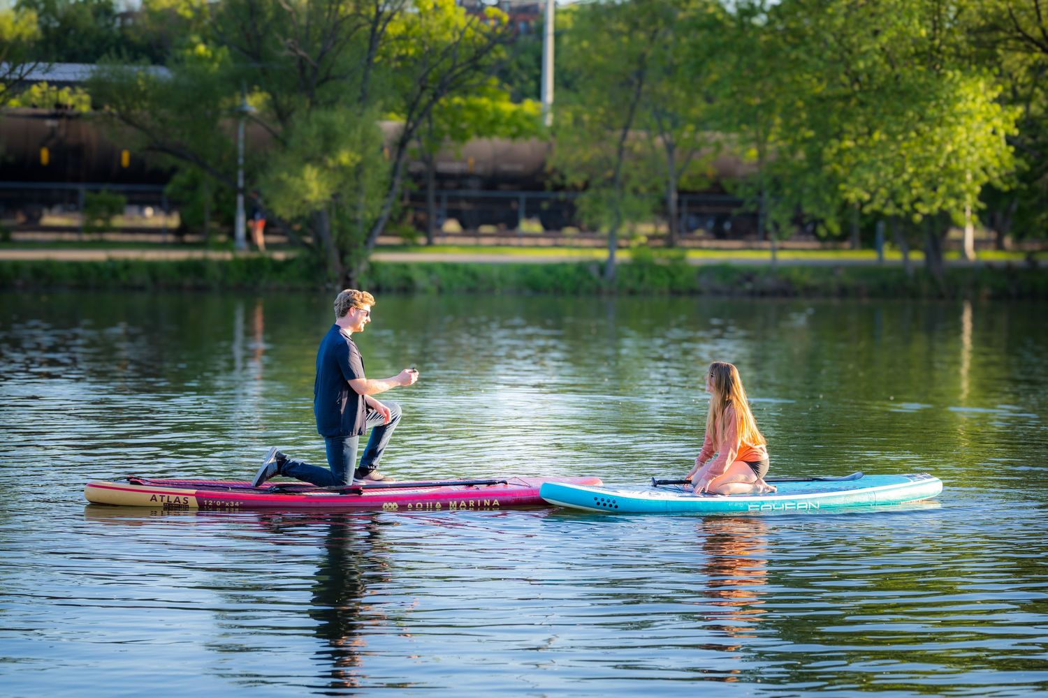 Two people on separate paddleboards on a calm lake surrounded by green trees in sunlight.