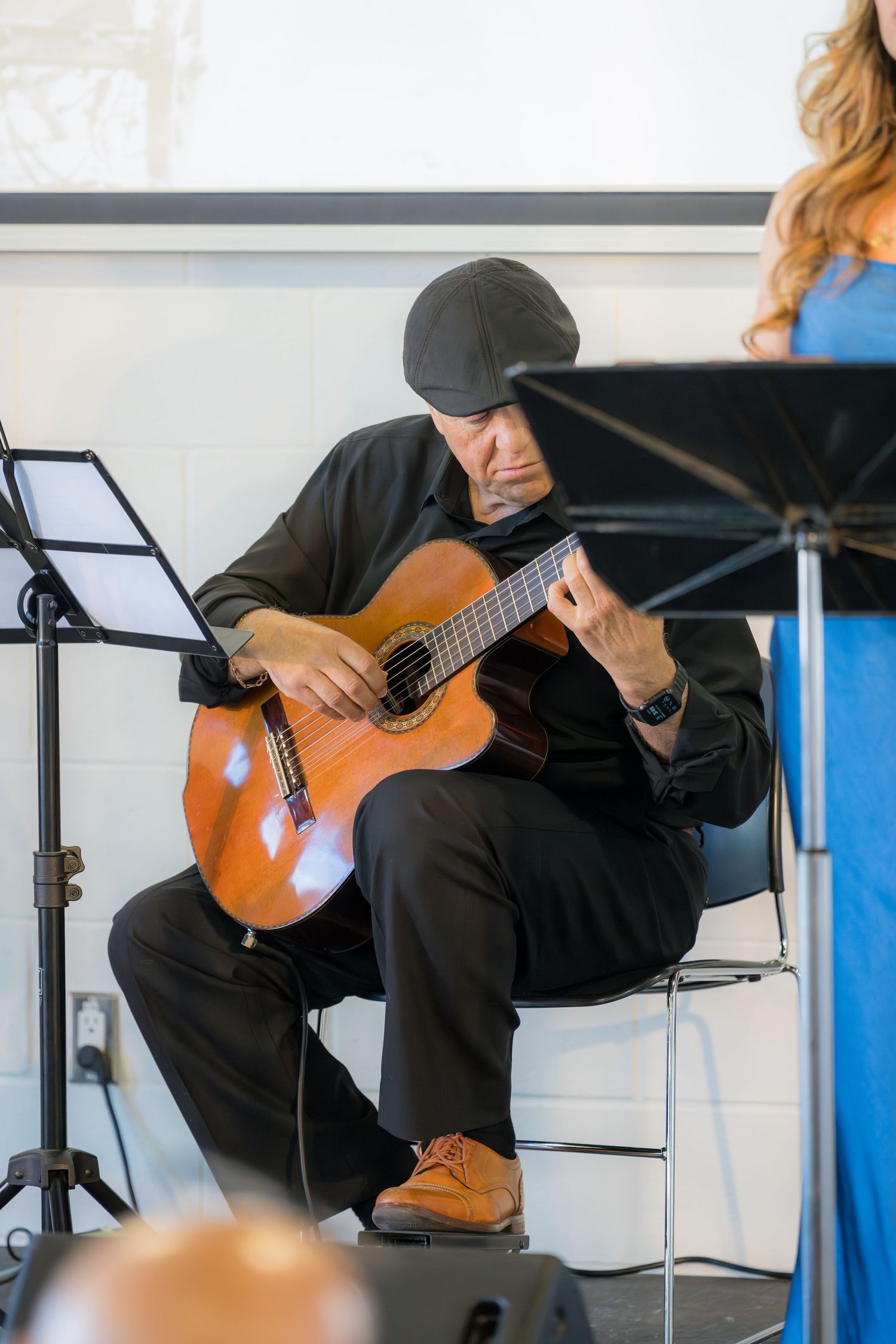 Male guitarist in black outfit and cap playing acoustic guitar at indoor event.