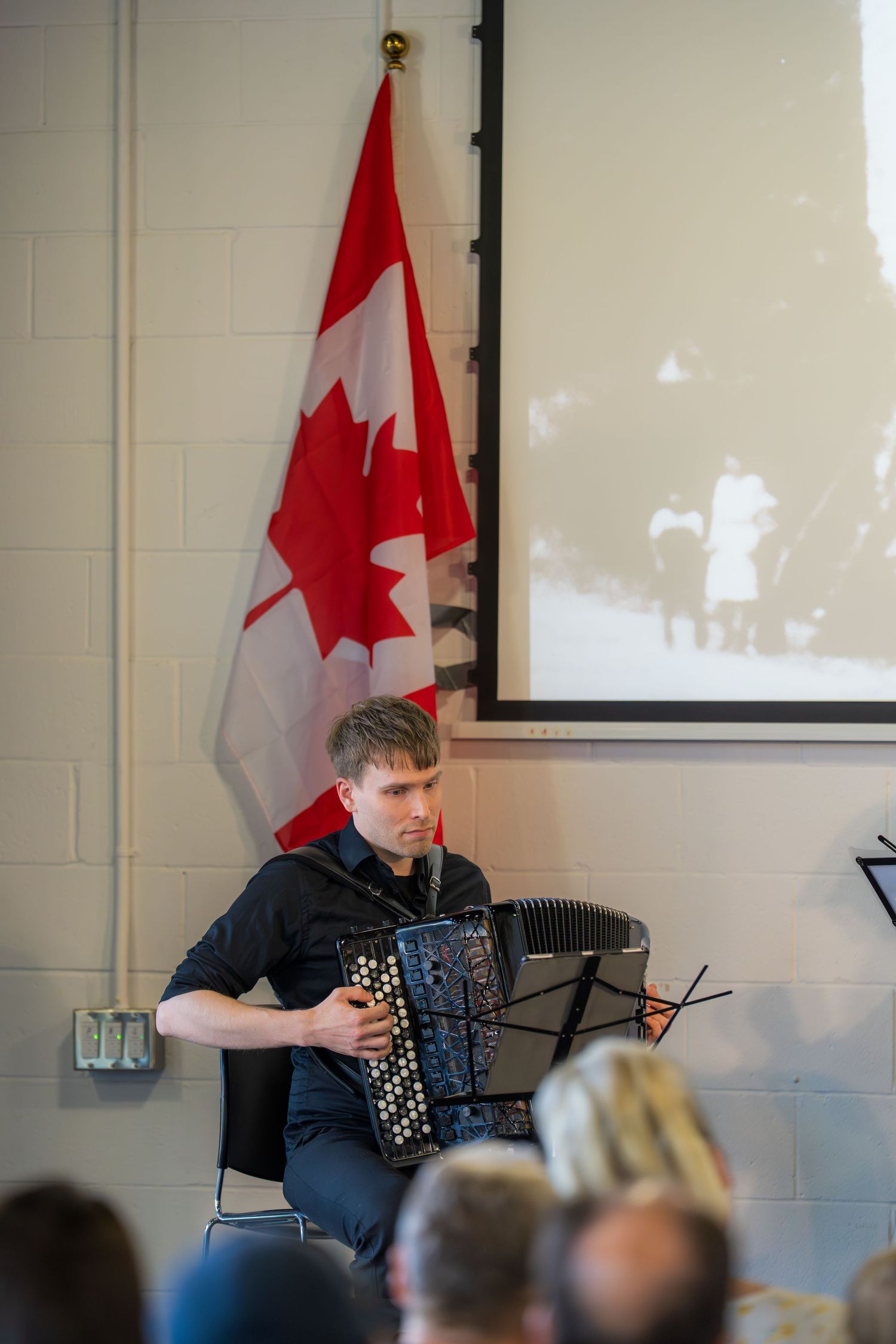 Musician playing accordion at Canadian event with maple leaf flag and historic photo projection in background.