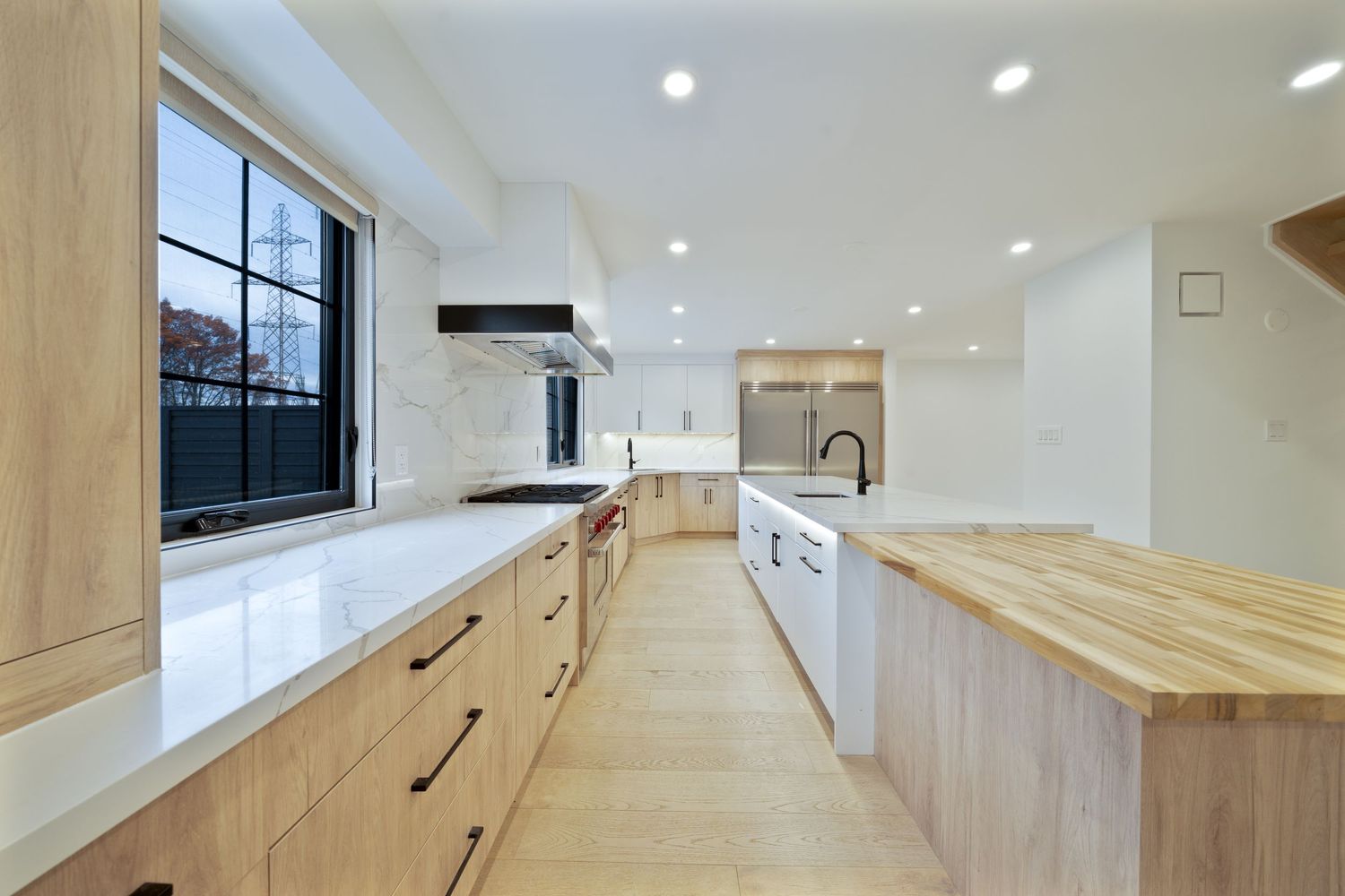 Modern kitchen with light wood cabinets, white countertops, and butcher block island in a bright open space with black windows.