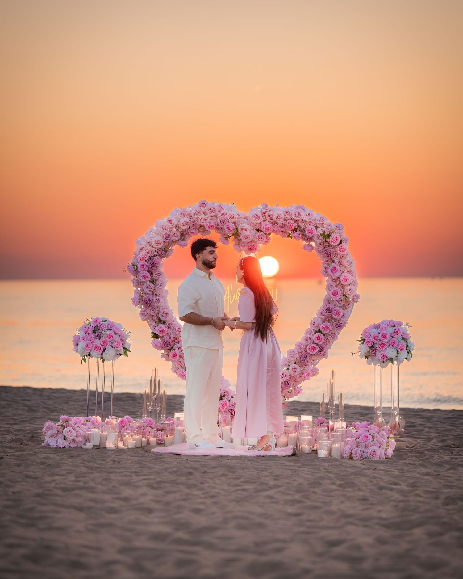 A romantic beach proposal setup with a heart-shaped pink floral arch against a stunning orange sunset.