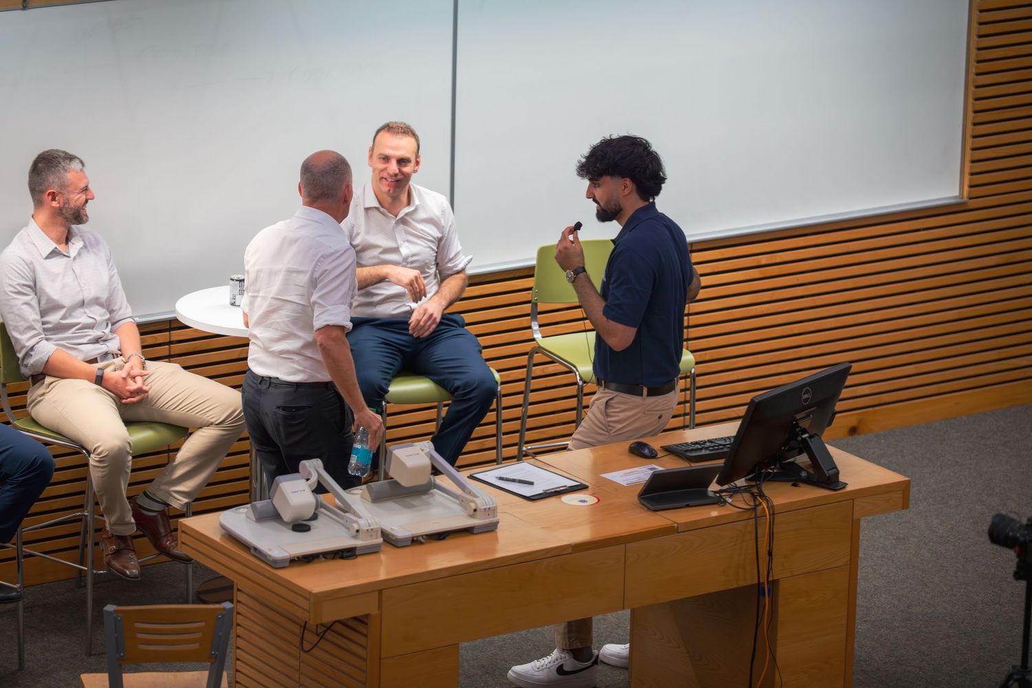 Students seated in tiered rows of desks in a modern lecture hall with wood paneled walls.