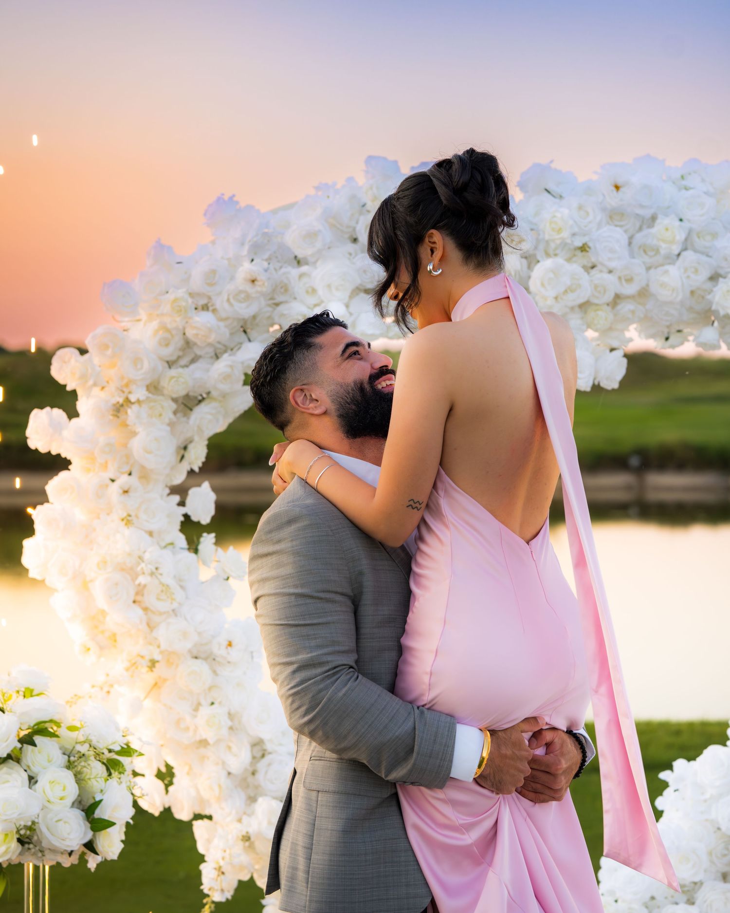 A couple shares a romantic kiss under a white floral arch during sunset on a golf course.