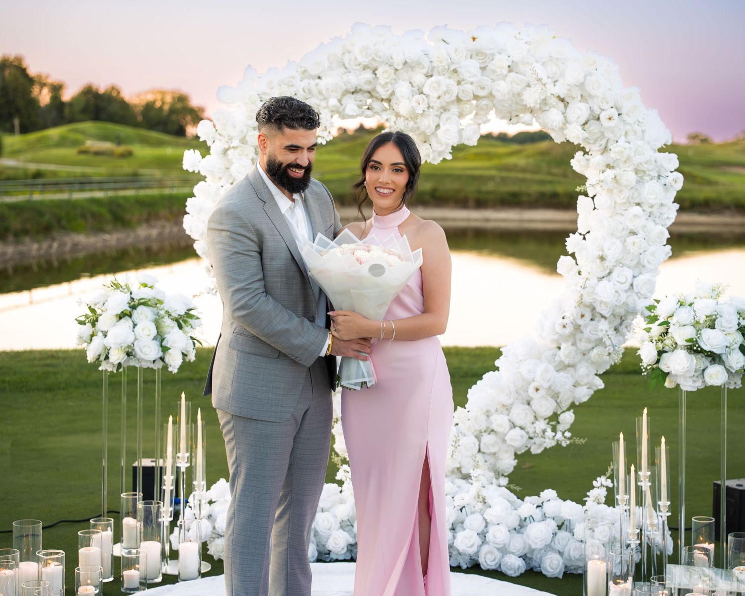A romantic proposal setup on a golf course with white floral arch and candles at sunset.