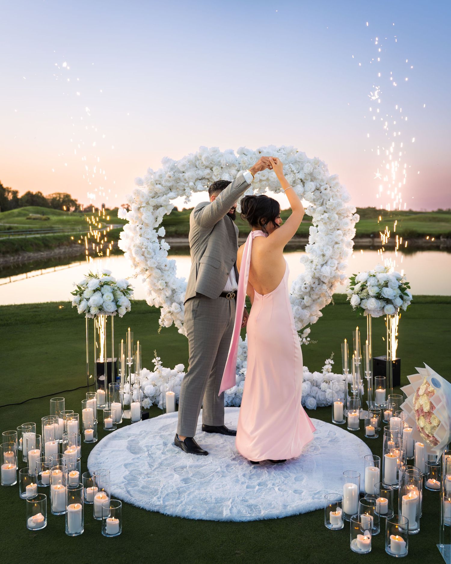 Elegant wedding ceremony setup with white floral arch and illuminated lanterns on a golf course.