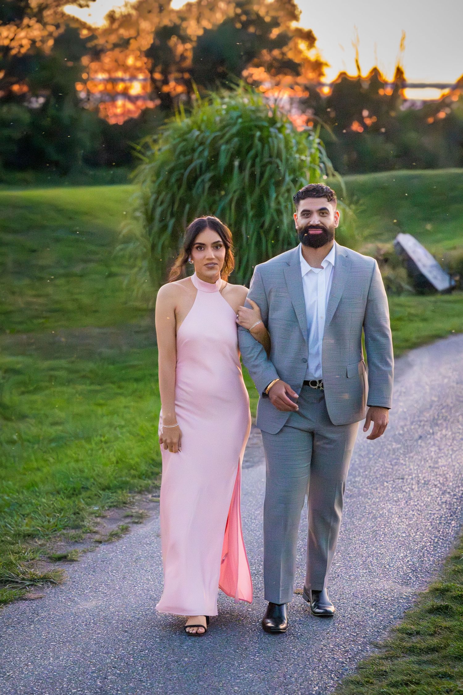 Two people stroll arm in arm along a scenic path at dusk wearing coordinated formal outfits.