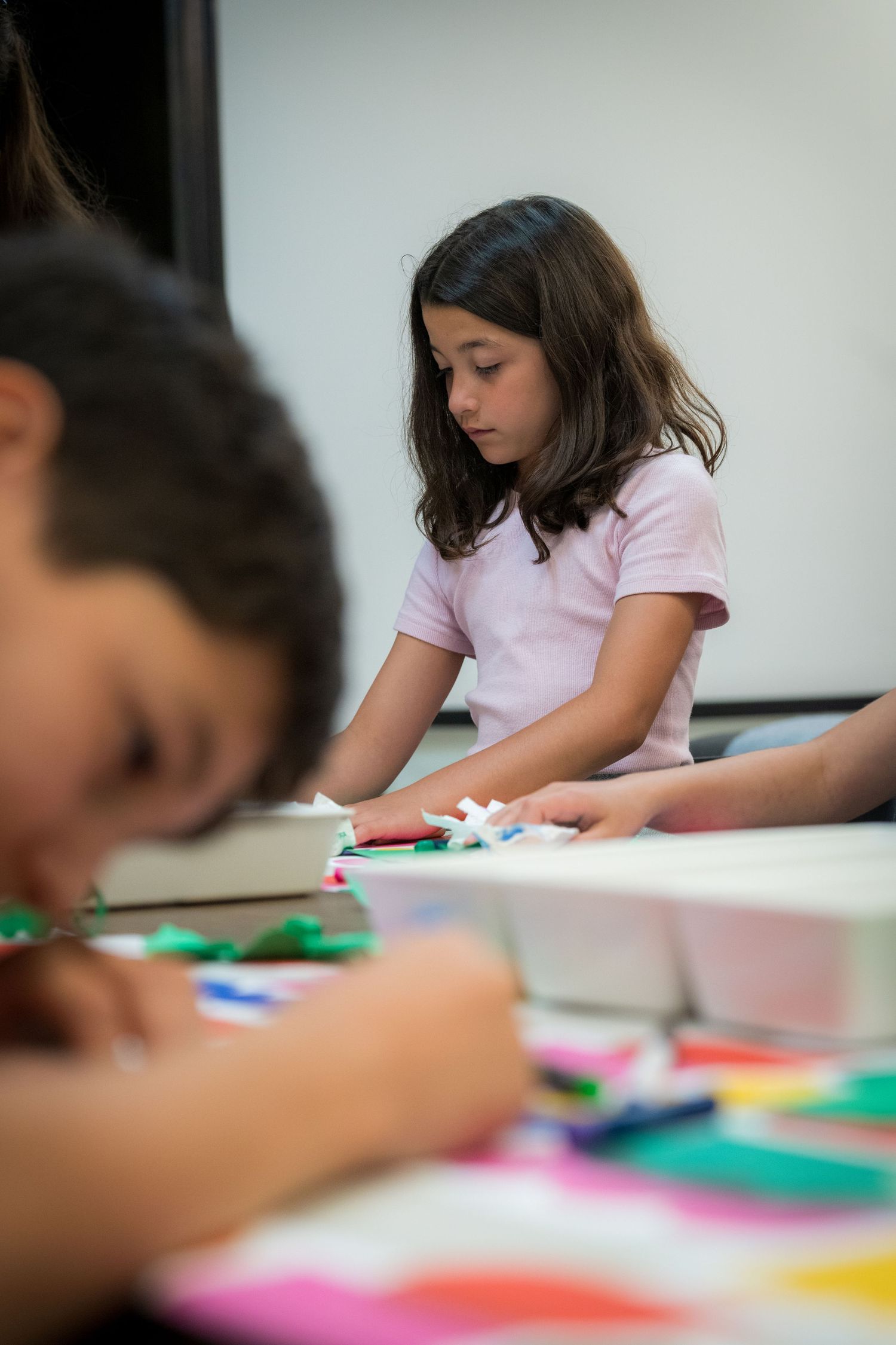 Girl in pink shirt carefully working on a bright paper collage art project at a table.