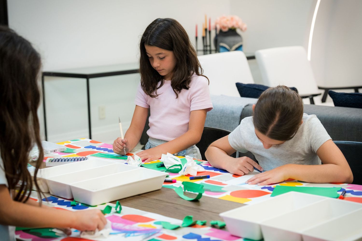 Three girls engage in a colorful paper craft activity together at an indoor table.