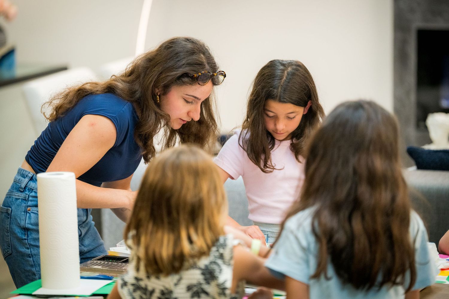 A teacher helps young girls with an art project at a colorful table indoors.