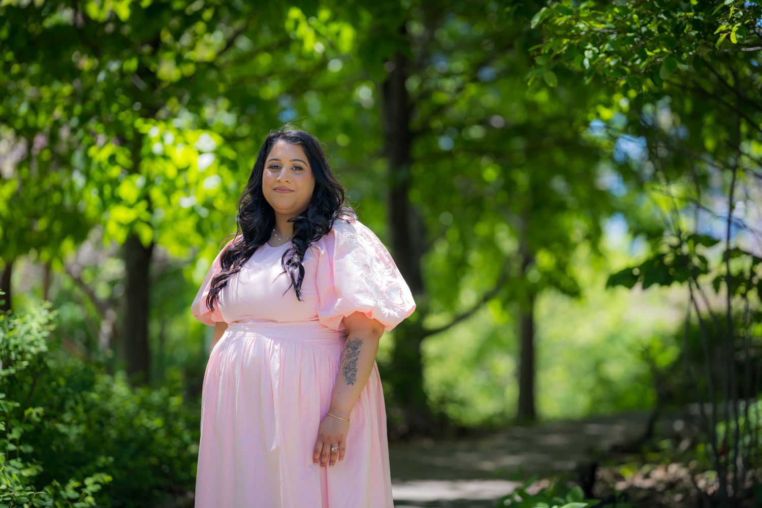 A pregnant woman in a flowing pink dress stands gracefully among lush green trees outdoors.
