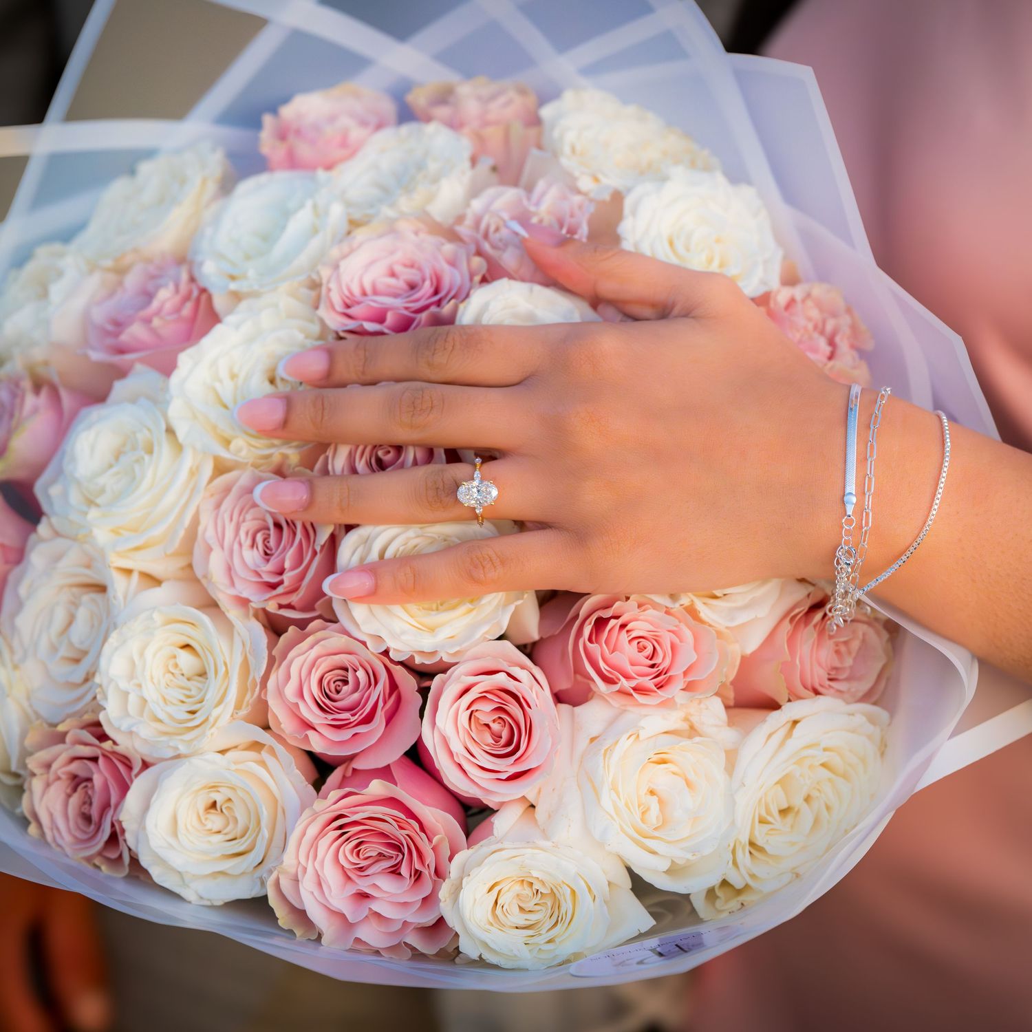 Close-up of hands holding a bouquet of pink and white roses wrapped in translucent paper.