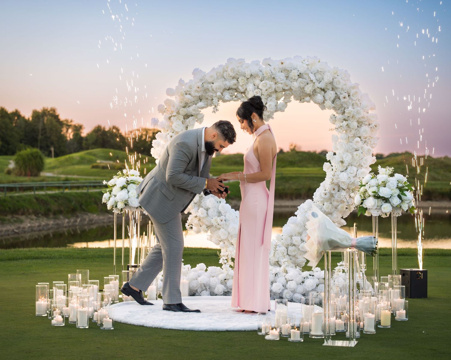 Beautiful outdoor proposal moment under white floral arch with glowing candles.