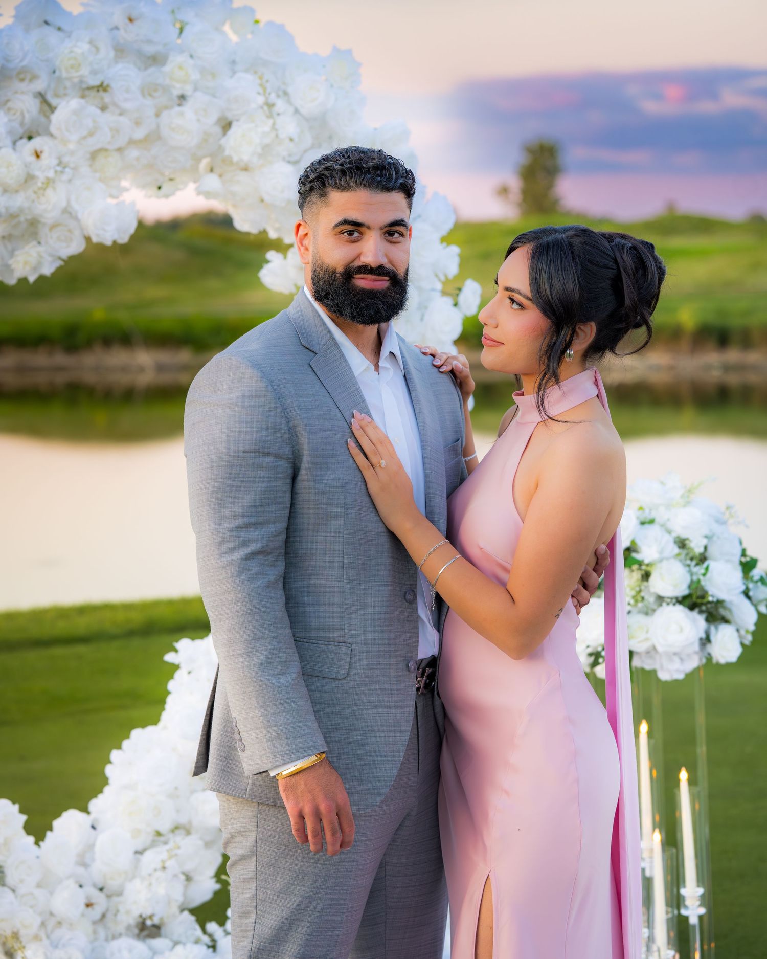 A couple shares loving glances under an elegant white floral arch at sunset.