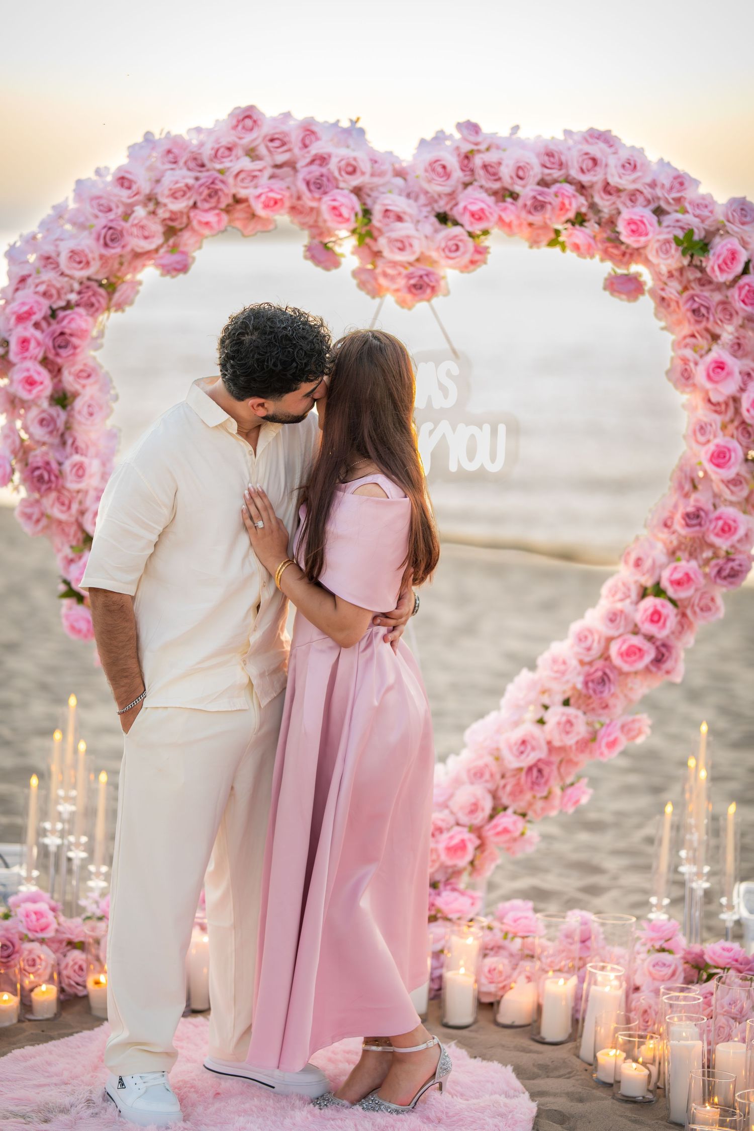 Pink rose heart-shaped arch with candles creates romantic beach proposal setting at sunset.