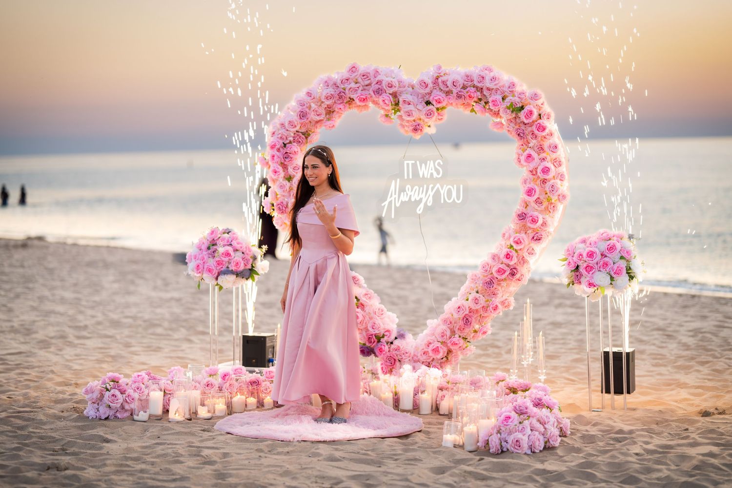 A romantic beach proposal scene with a pink floral heart arch and sparkling lights at dusk.