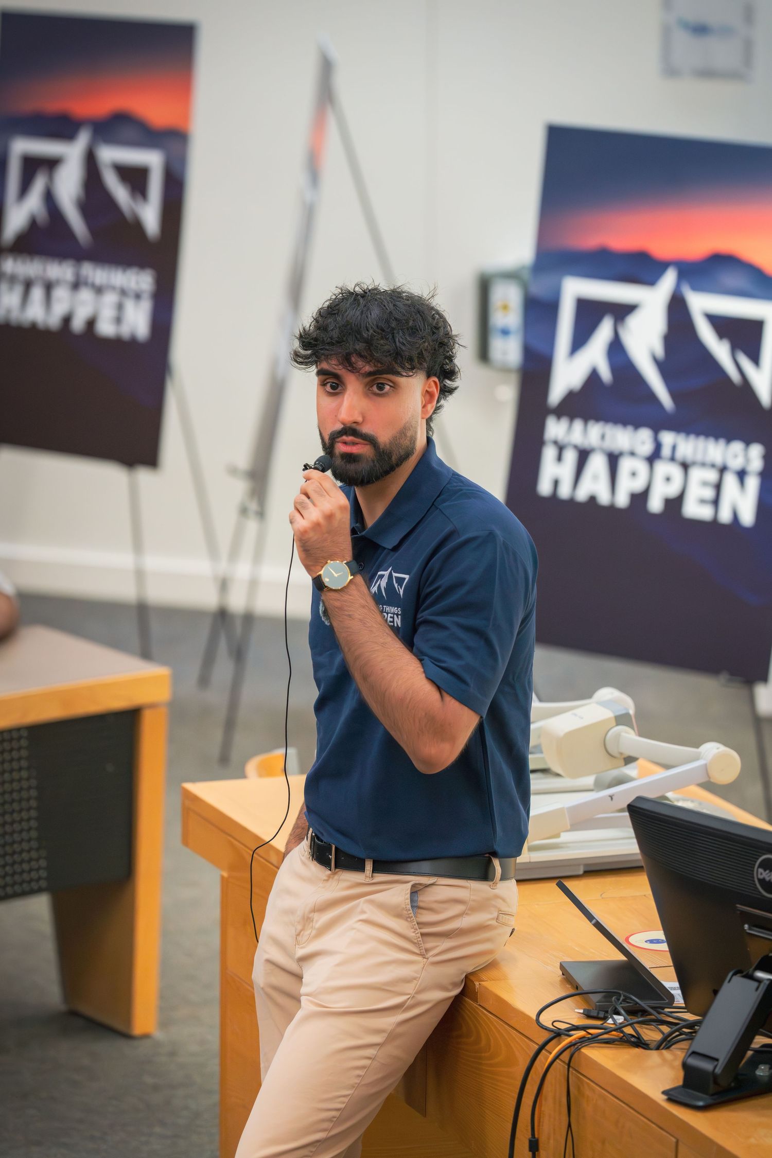 Person in a navy polo and khaki pants gesturing while seated at a desk with a computer monitor.
