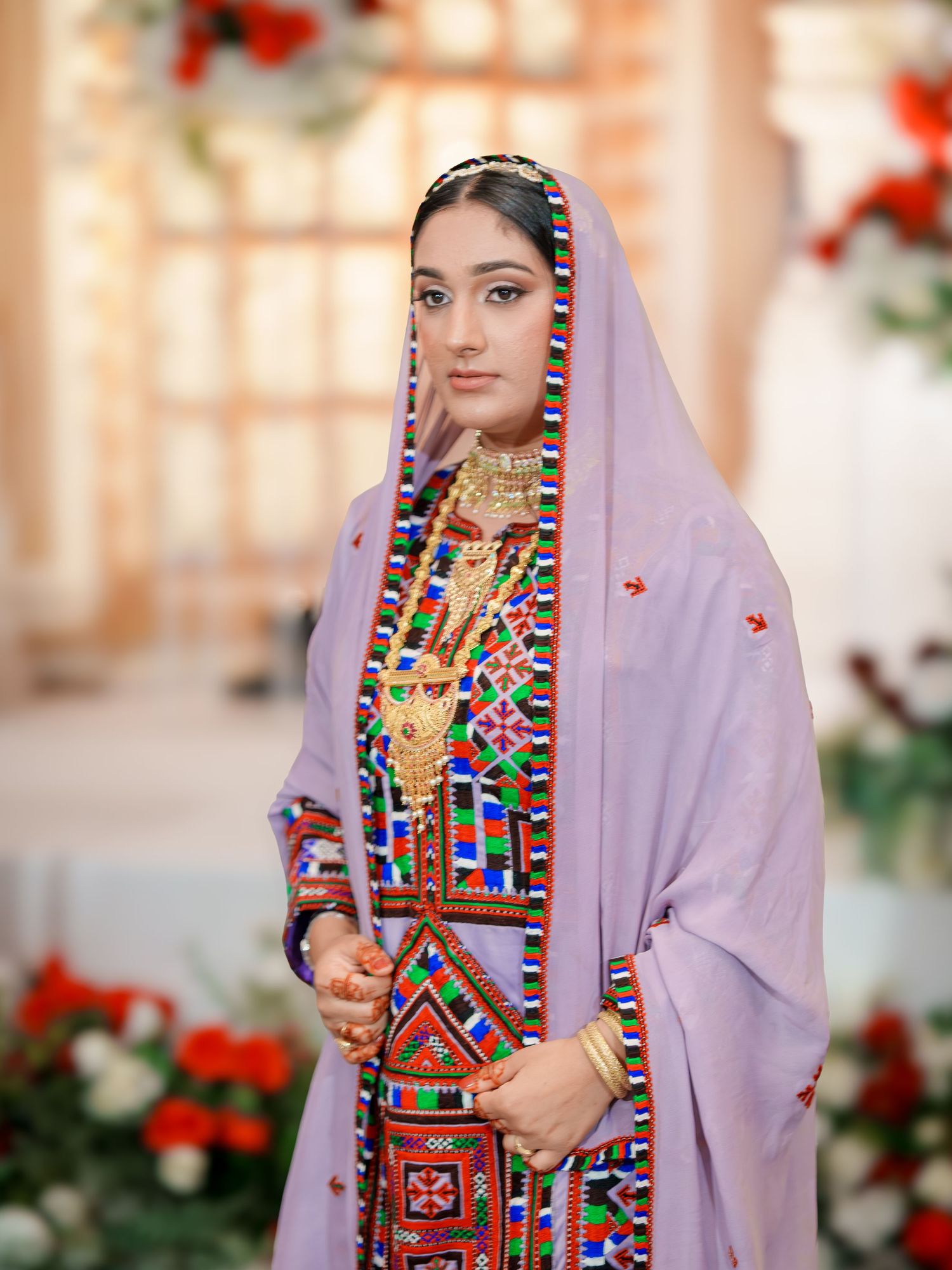 Woman in traditional Afghan dress with colorful geometric embroidery and lavender veil.