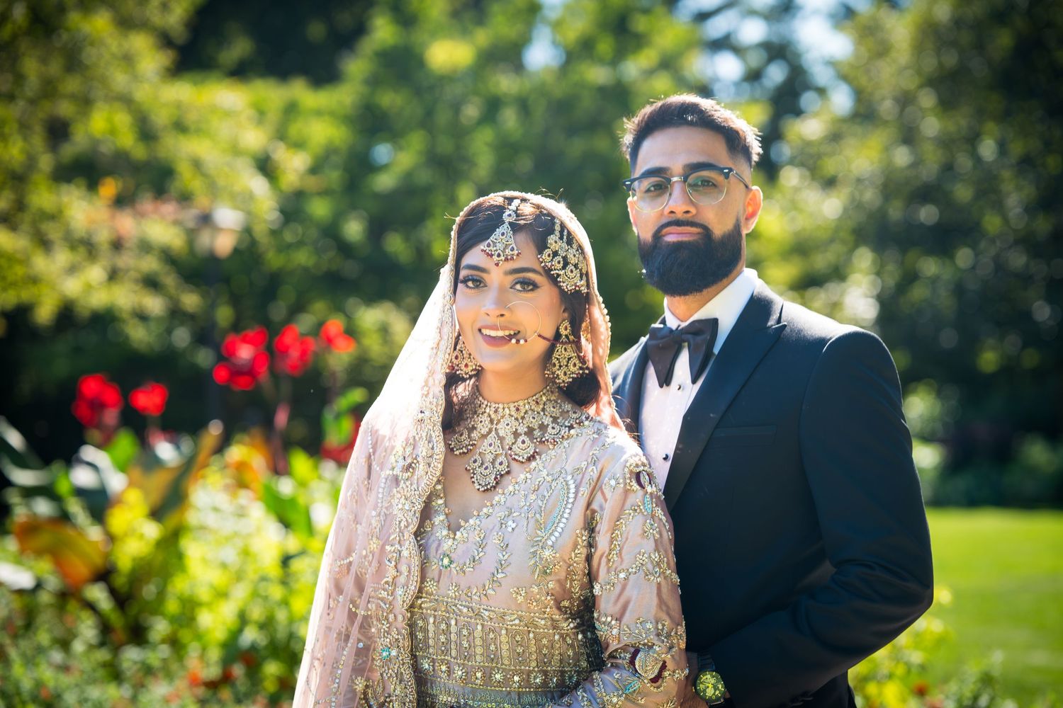 Traditional South Asian wedding couple poses in garden setting wearing ornate bridal attire and formal suit.