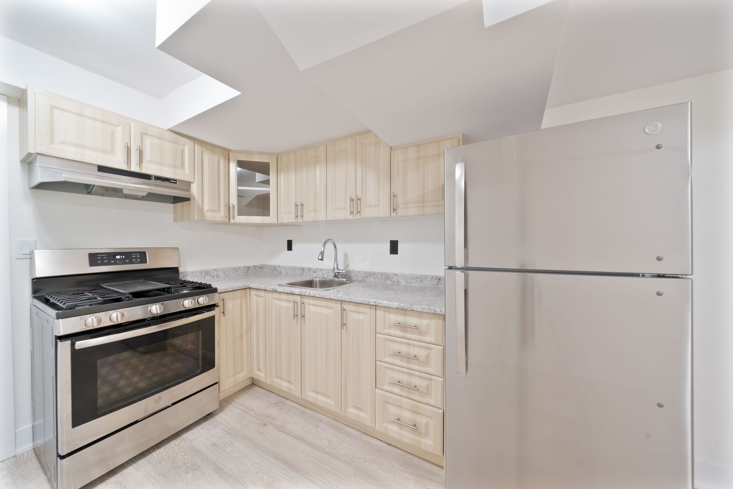 Modern L-shaped kitchen with light wood cabinets, stainless steel appliances, and white marble countertops in a bright space.
