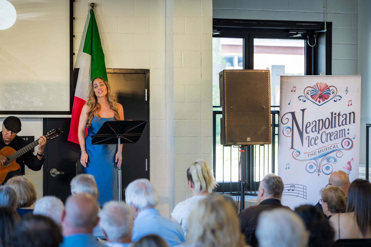 Female vocalist in blue dress performs at Neapolitan Ice Cream The Musical with guitarist beside Italian flag.