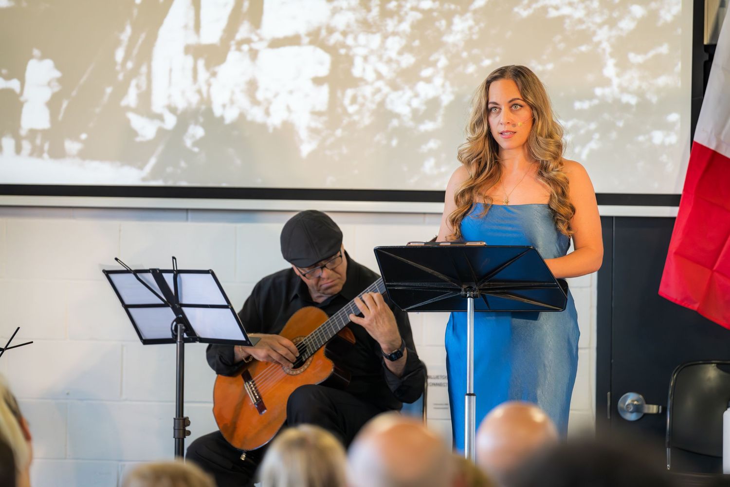 Woman in blue dress presents at podium while guitarist performs at Italian cultural event.