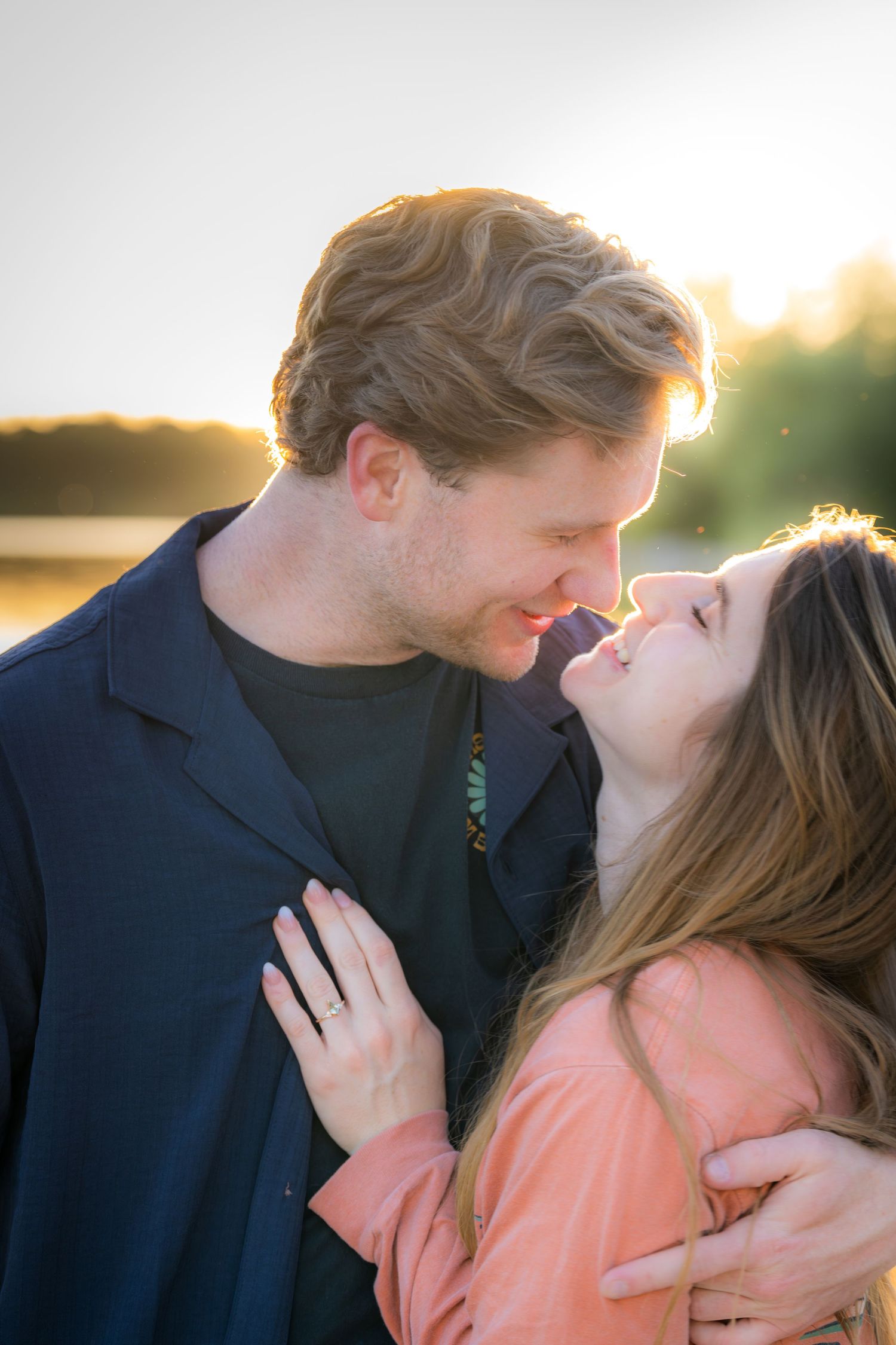 Couple sharing a romantic nose-to-nose moment outdoors at golden hour, woman wearing engagement ring.