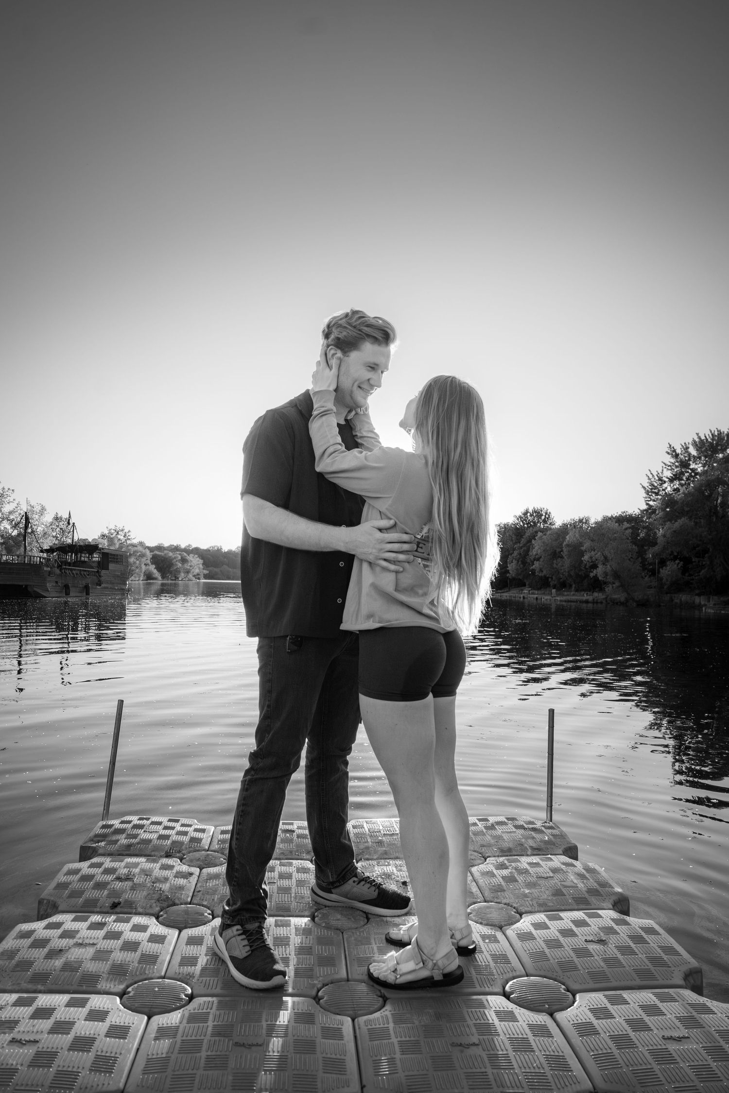 Black and white photo of a couple embracing on a floating dock by a calm lake at sunset.