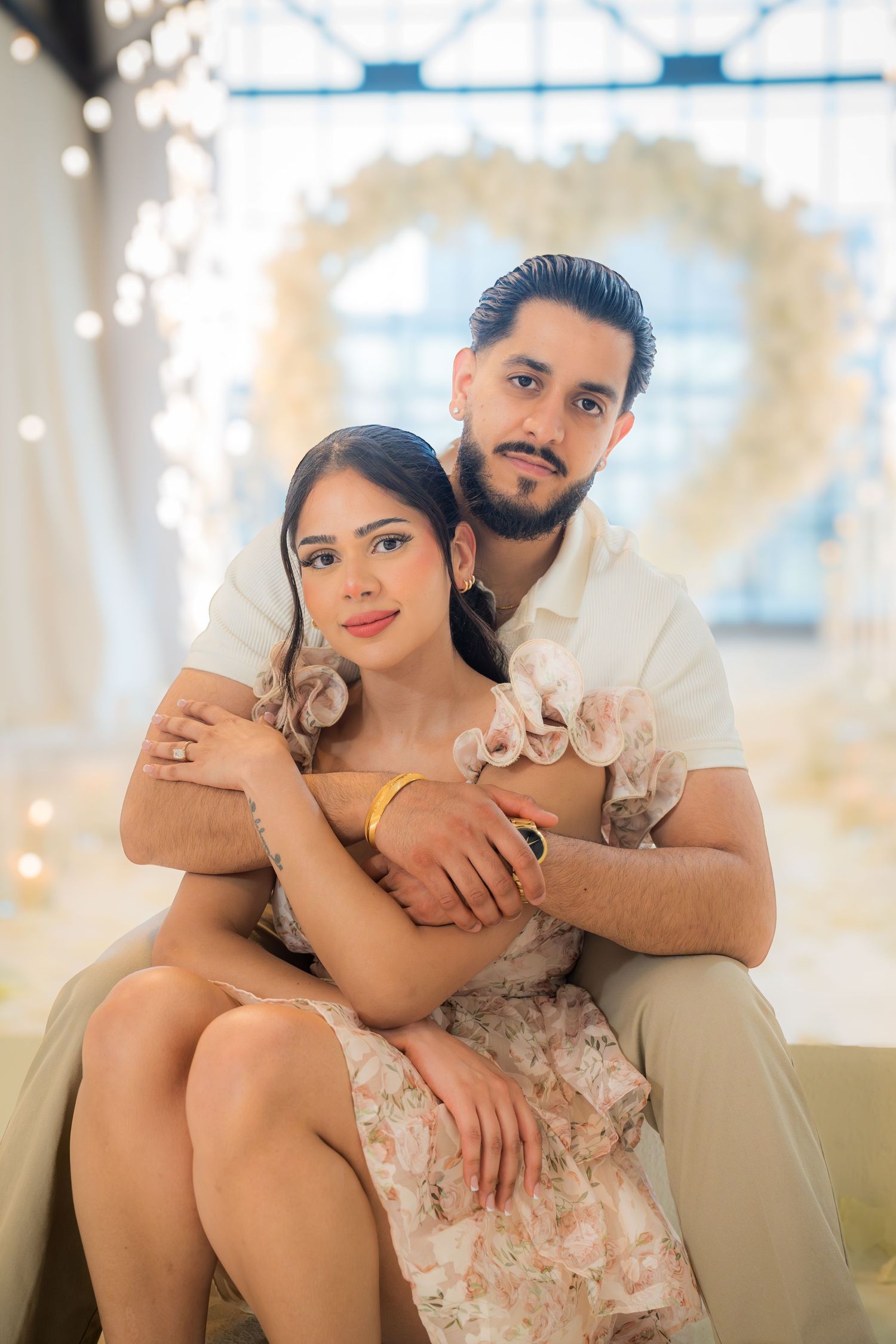 A happy engaged couple poses together in front of a glowing white floral heart arch backdrop.