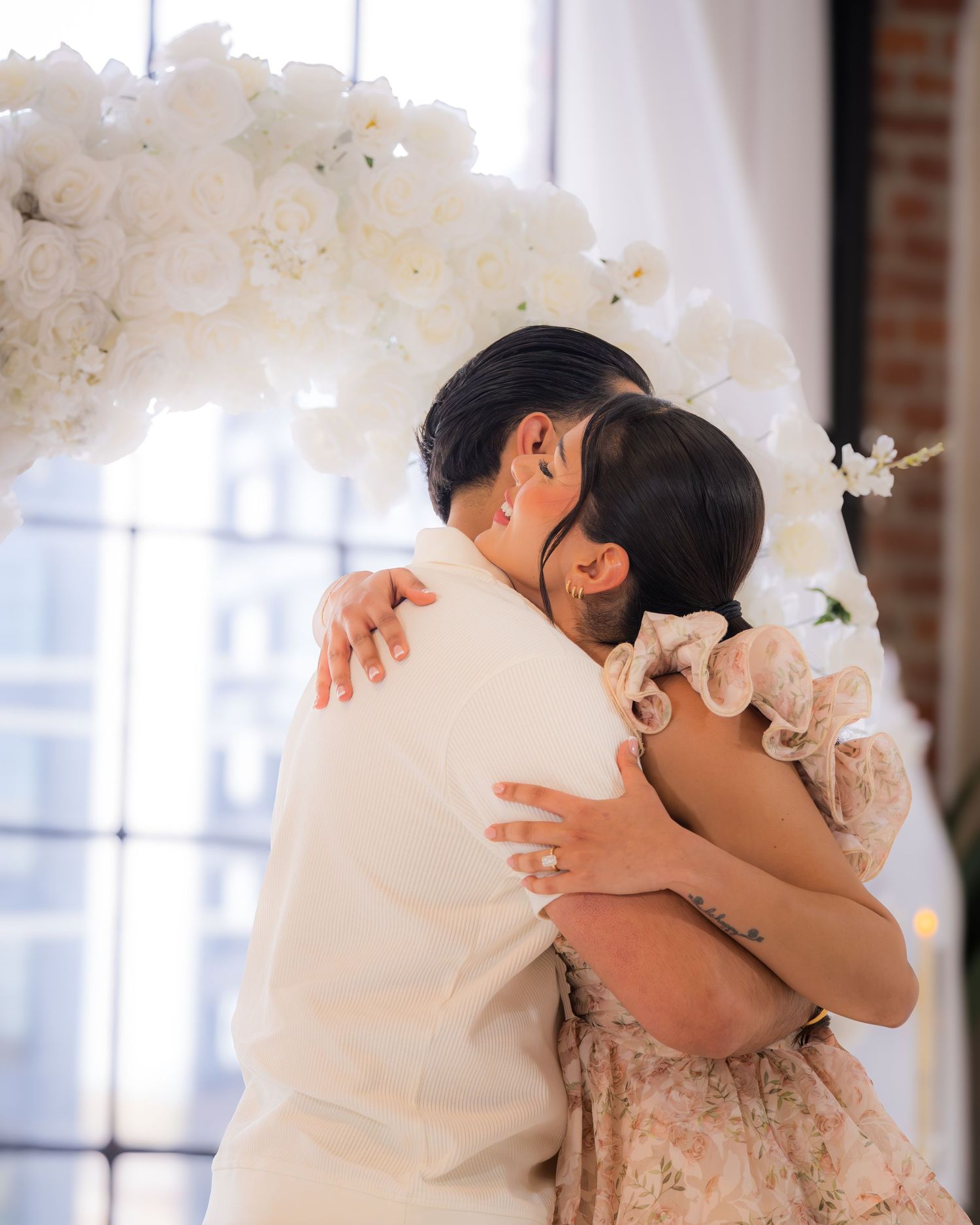 Couple sharing an emotional hug beneath a white floral arch during a romantic proposal moment.